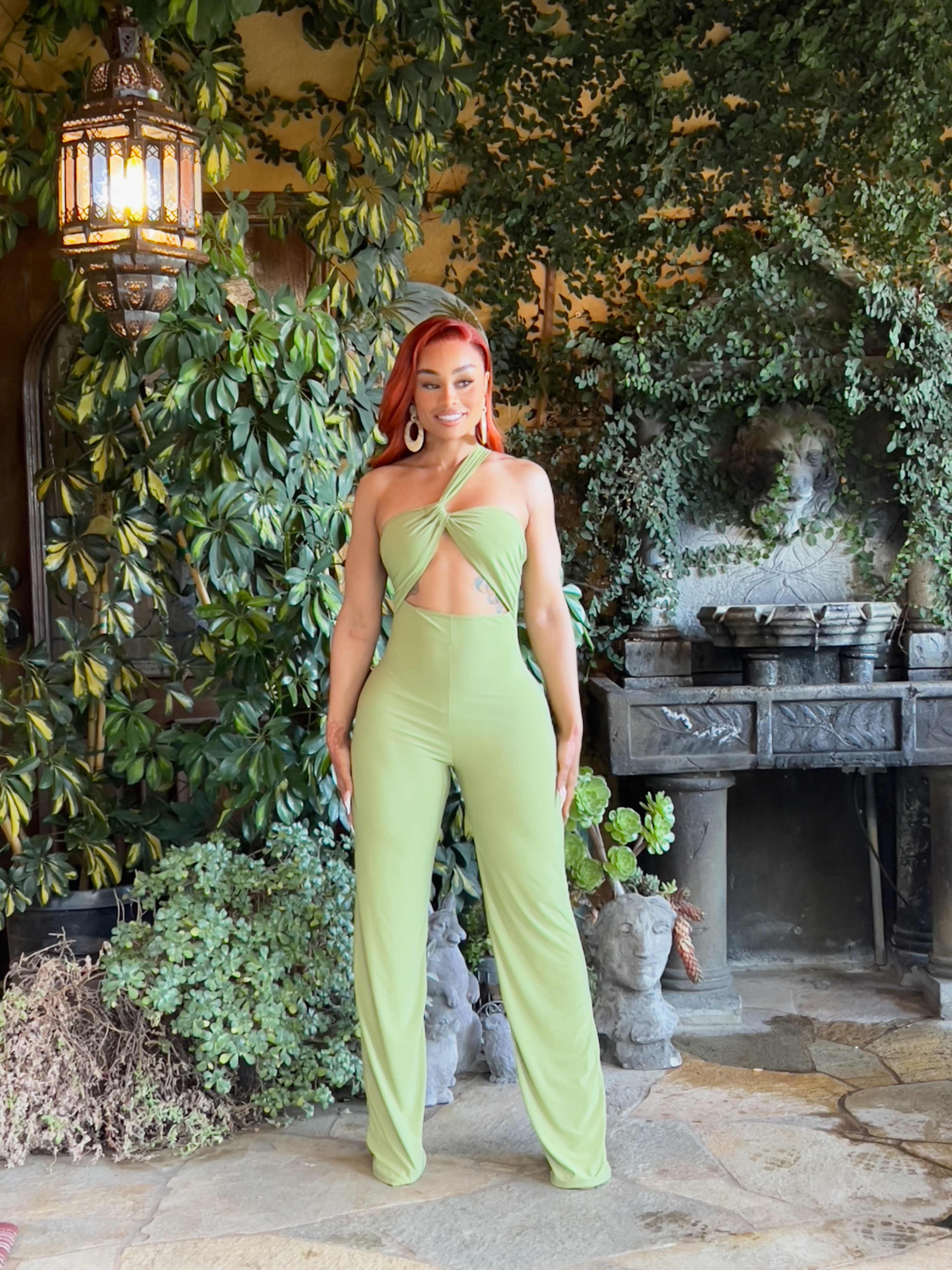 A person stands in front of a stone structure adorned with plants, wearing a green jumpsuit.