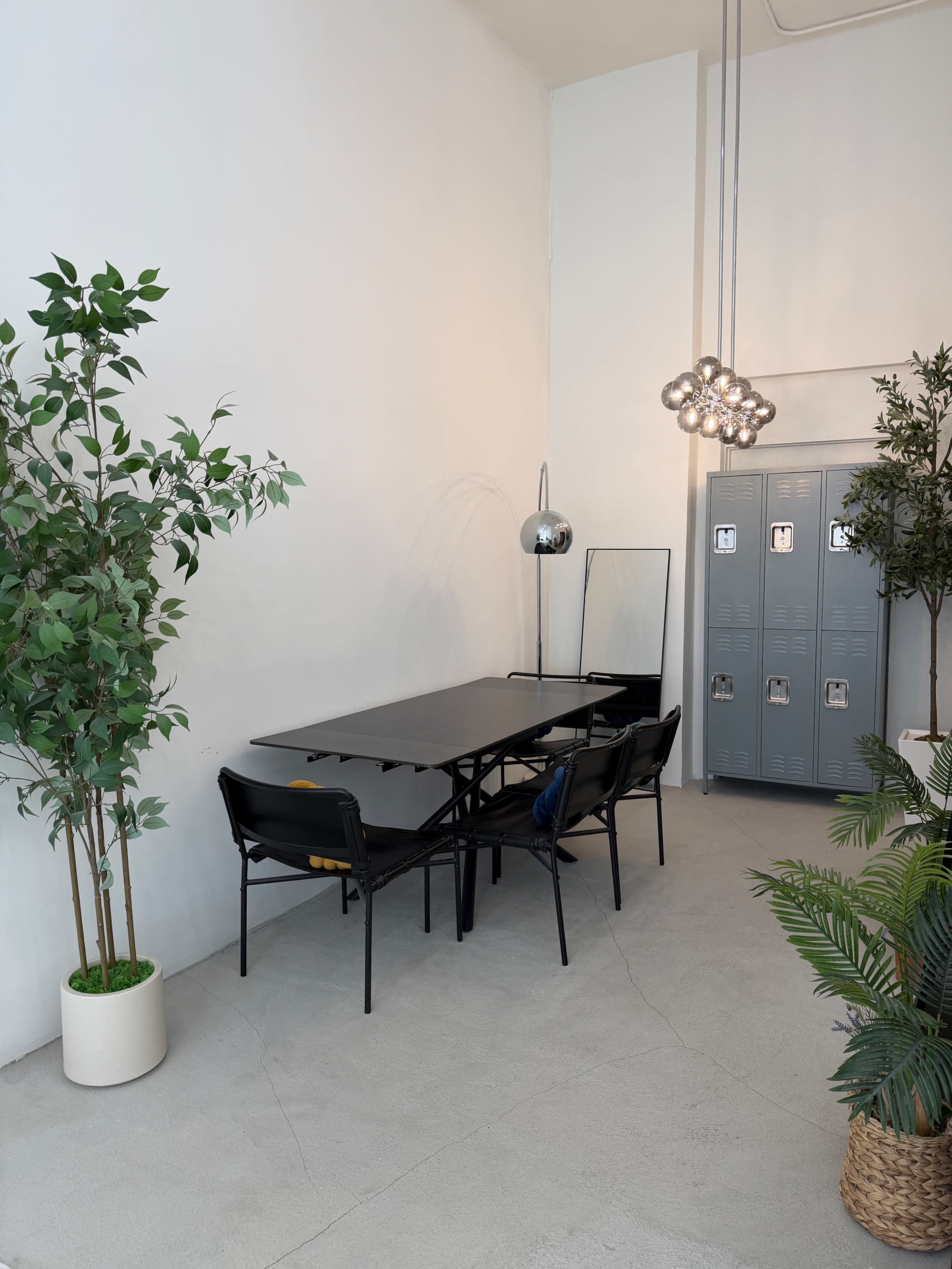 The image shows a minimalistic office space featuring a black table with chairs, a decorative plant, and gray lockers against a white wall.