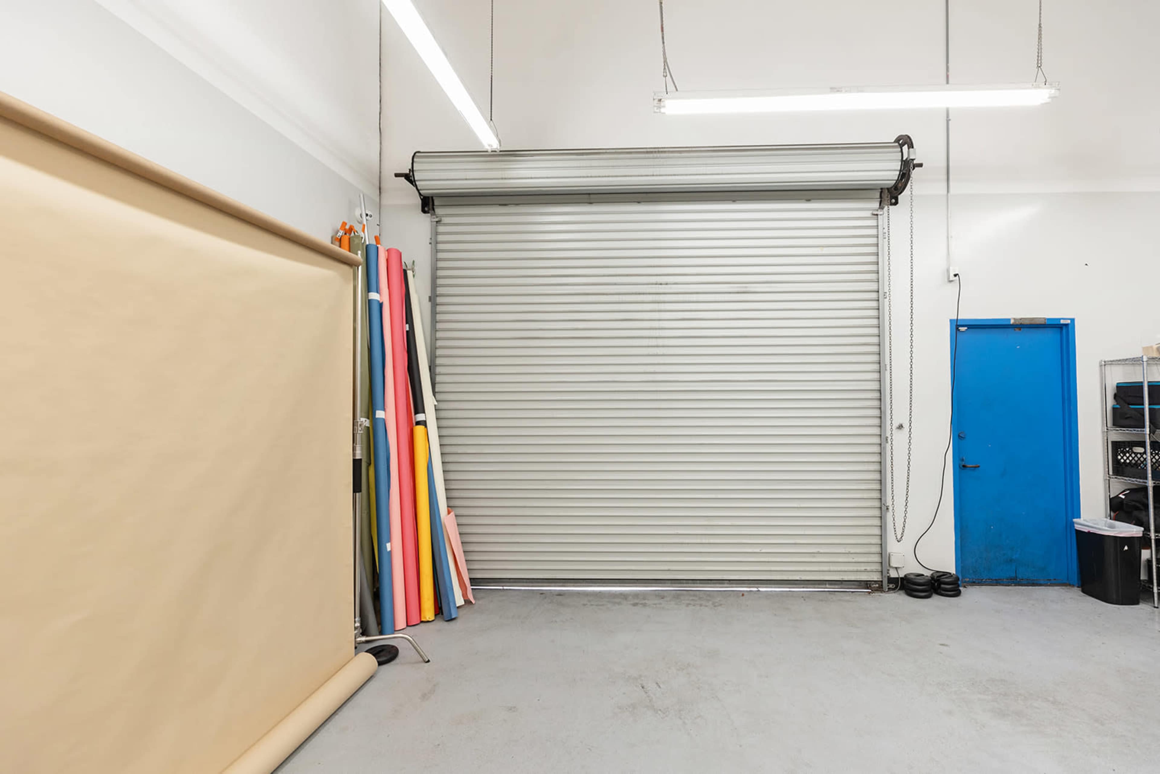 A storage space features a roll-up metal door, a blue door, and a backdrop of colorful paper rolls stacked against the wall.