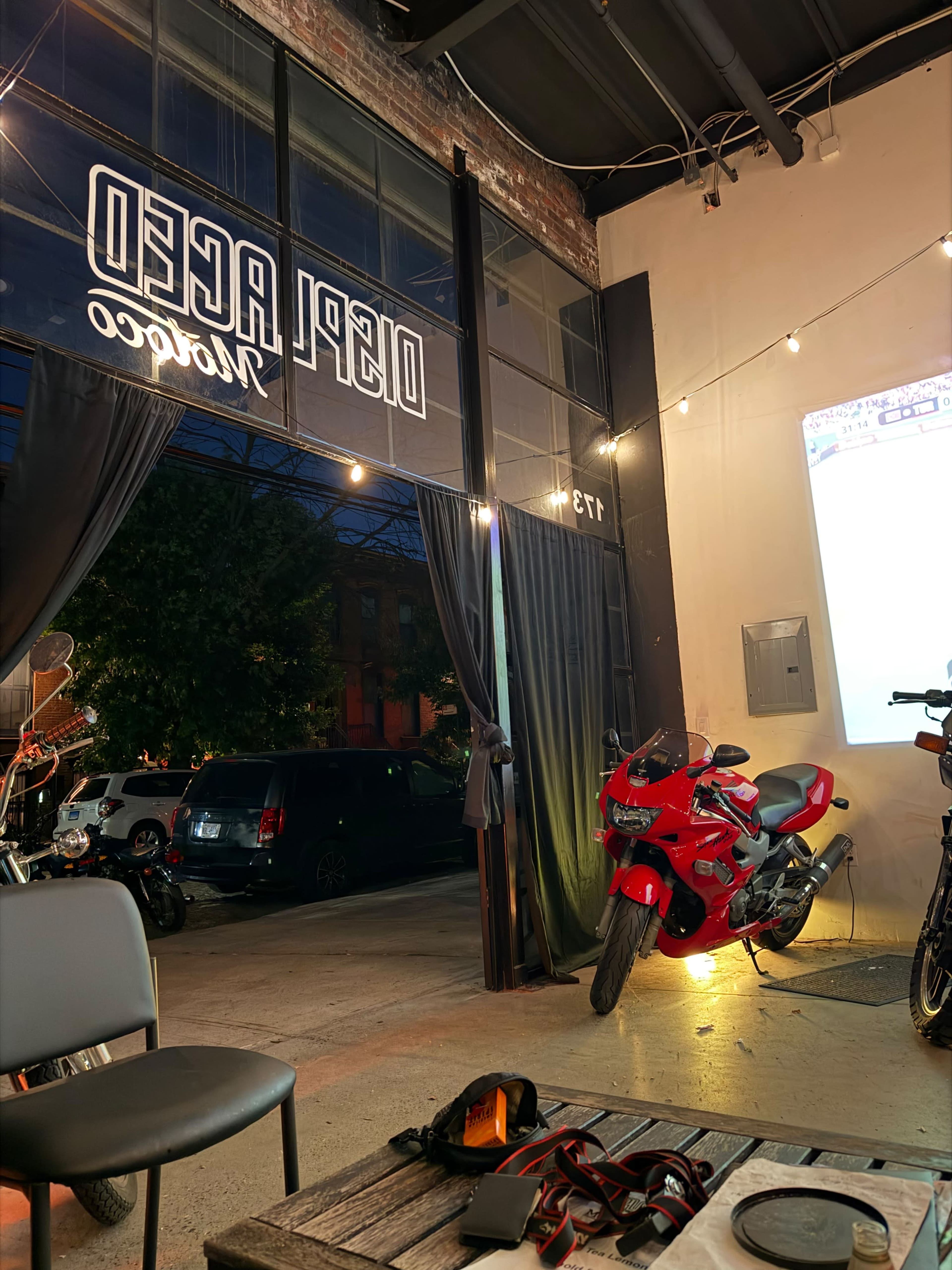 The image shows a dimly lit cafe or workspace featuring a large garage door opening, a red motorcycle, and string lights overhead.