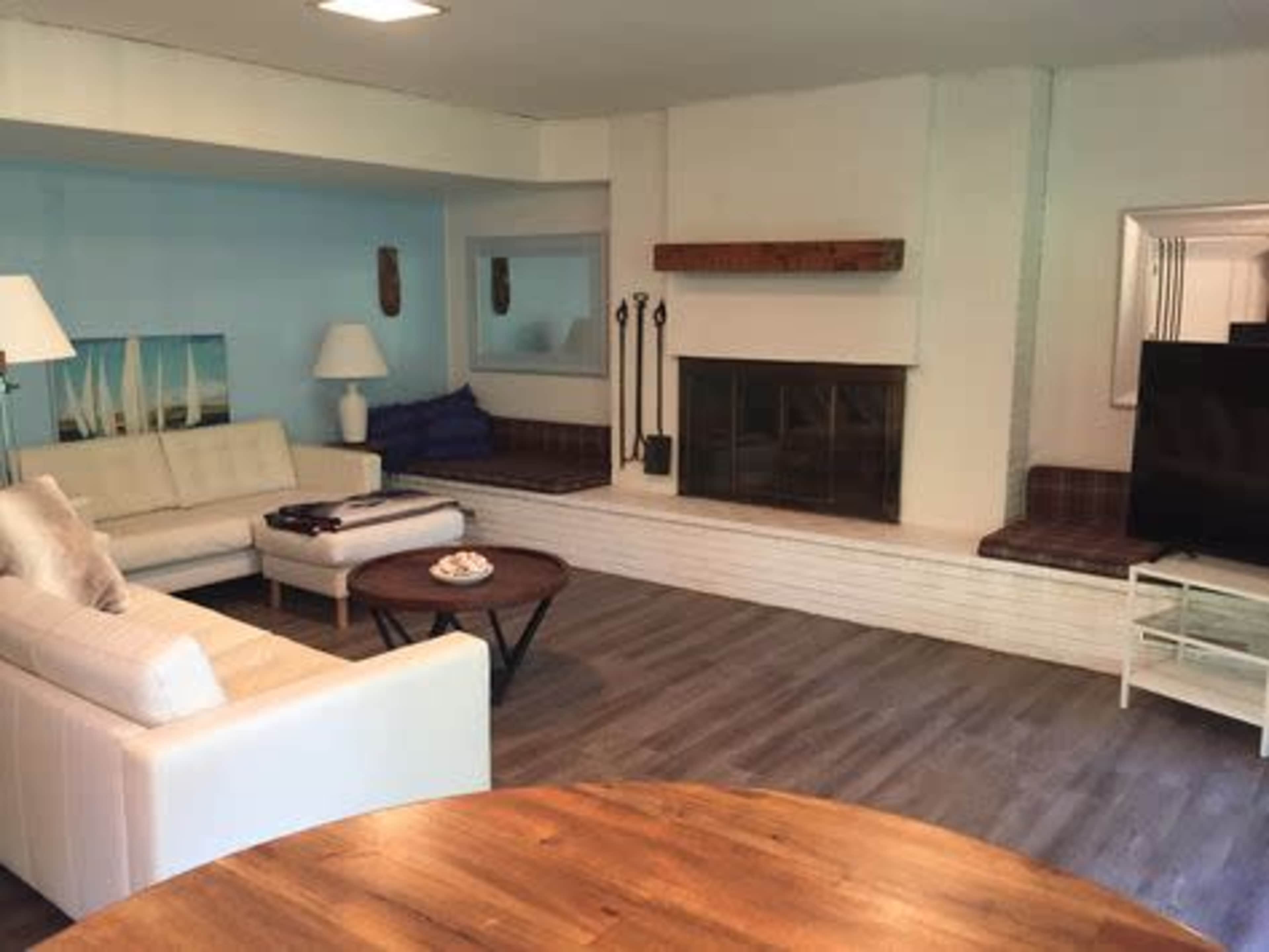 The image shows a modern living room featuring a white sofa, a round wooden coffee table, a television on a glass stand, and a fireplace with a wooden mantel.