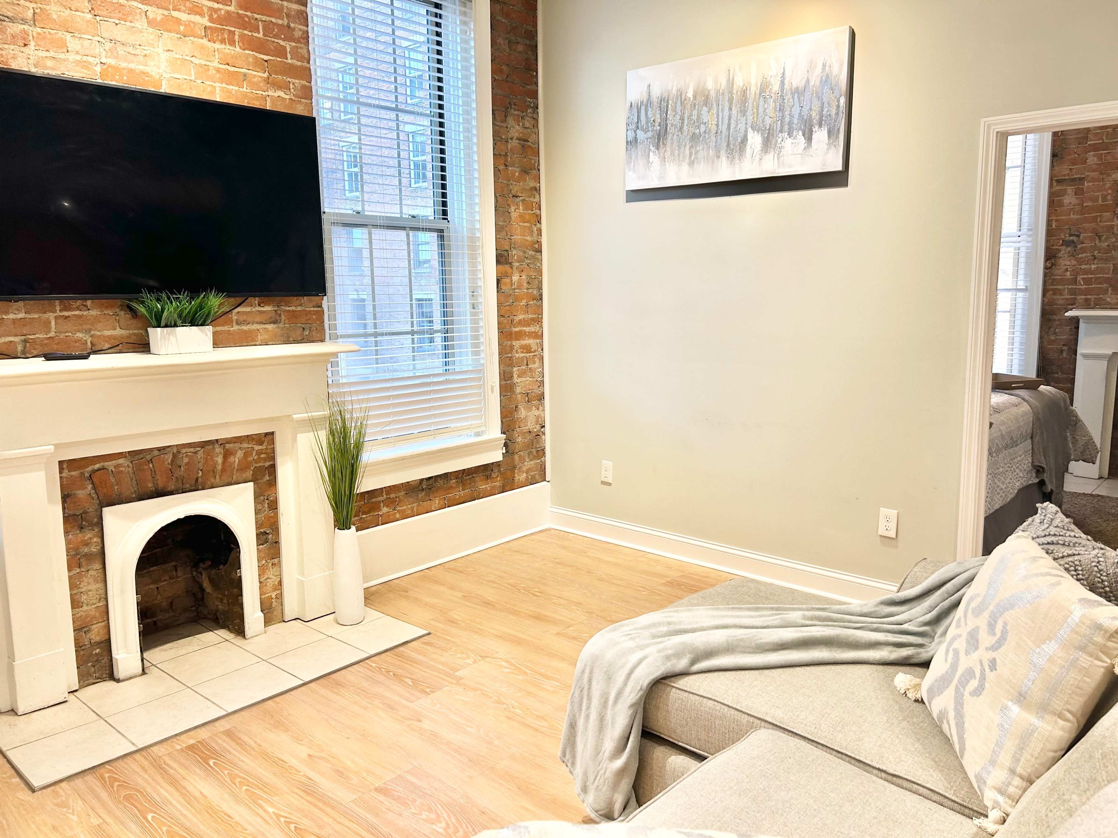A modern living room with a flat-screen TV mounted above a white fireplace, brick walls, a light-colored sofa, and a doorway leading to a bedroom.