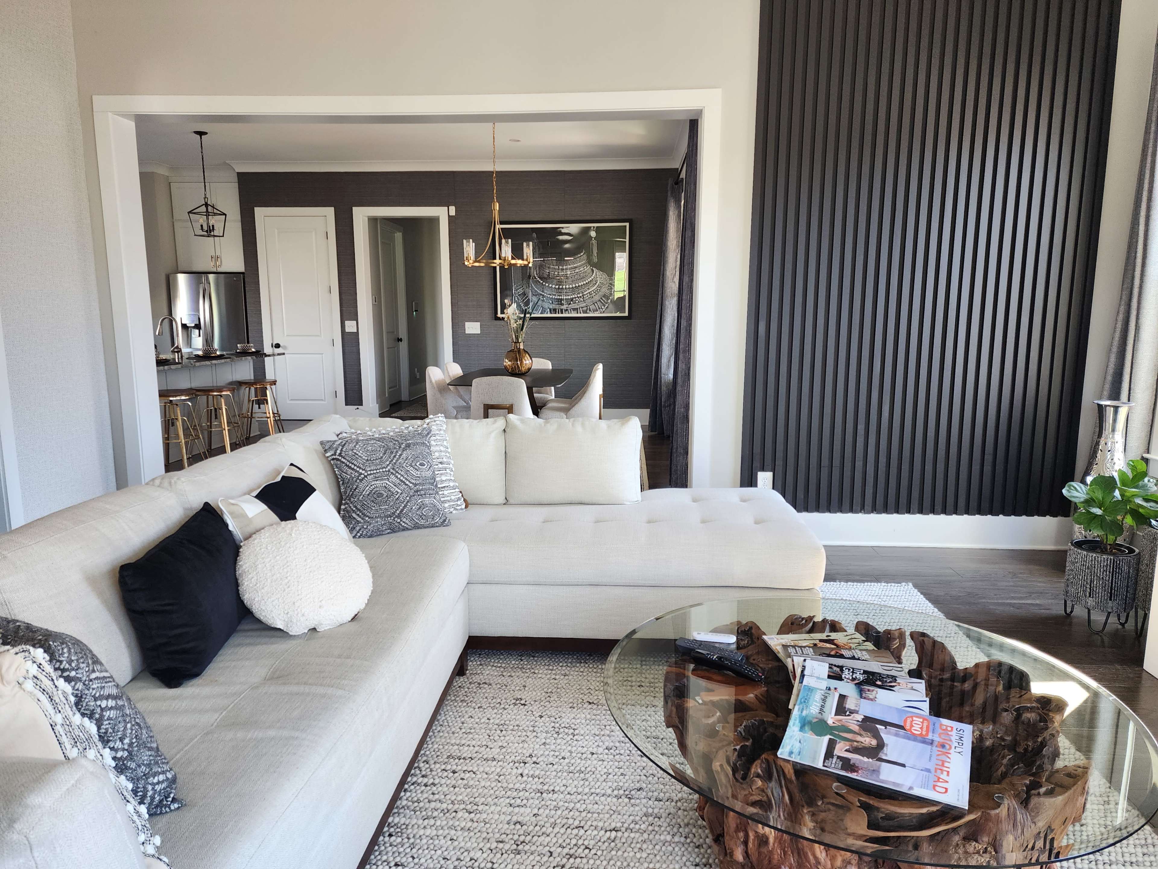 The image shows a modern living room with a light-colored sectional sofa, a glass coffee table with a wooden base, and a view into a dining area with a dark accent wall and contemporary decor.