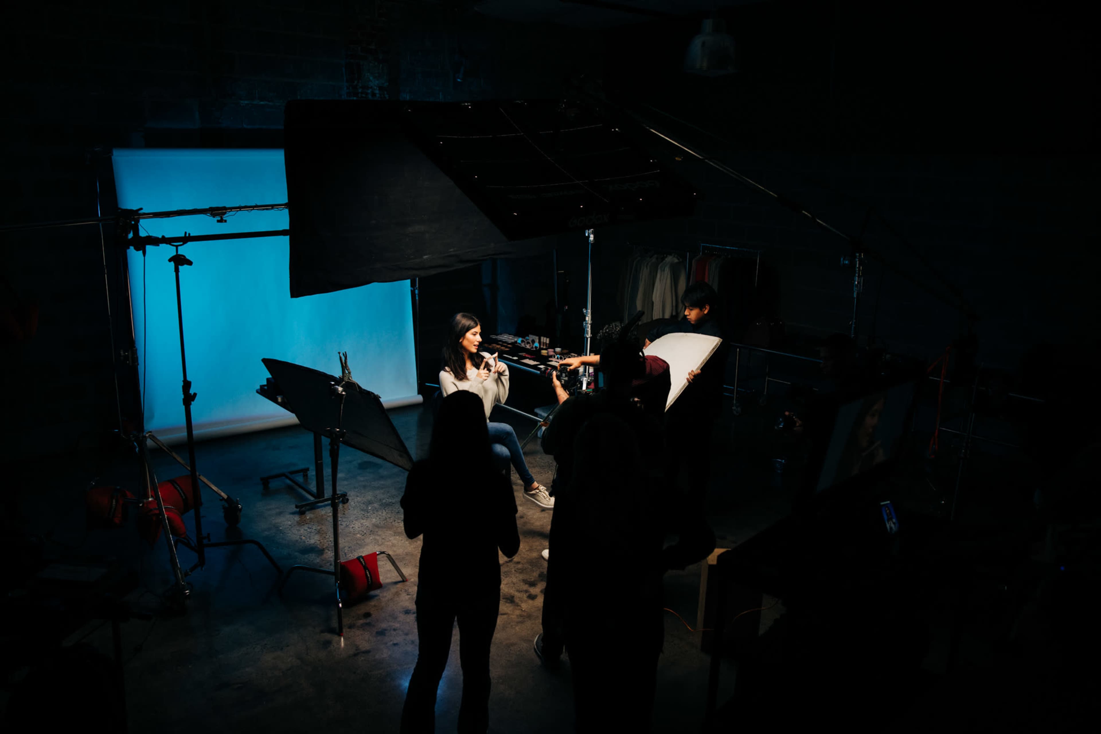 A model sits for a photoshoot in a dimly lit studio with a blue backdrop and several lighting equipment setups.