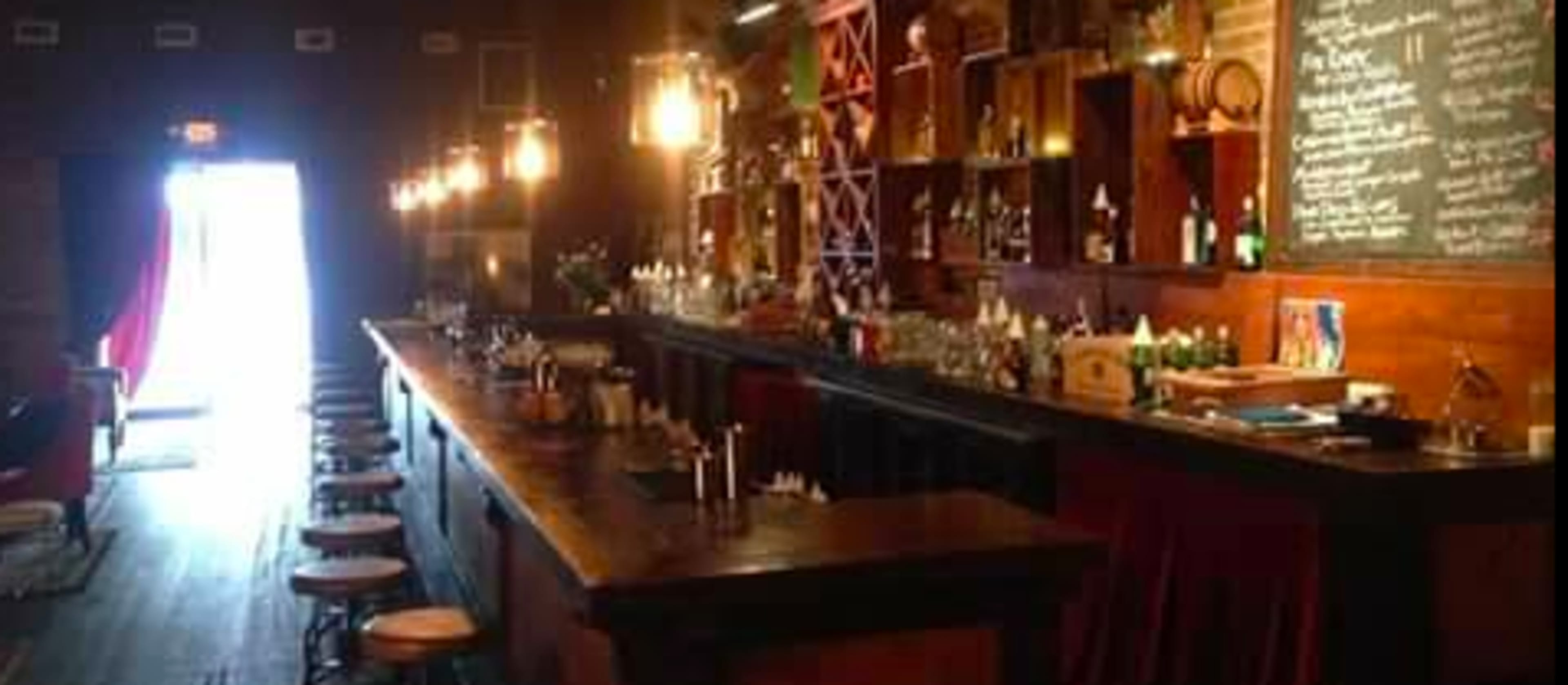 A long, wooden bar is lined with stools, illuminated by hanging lights, and features shelves stocked with various bottles against a brick wall.