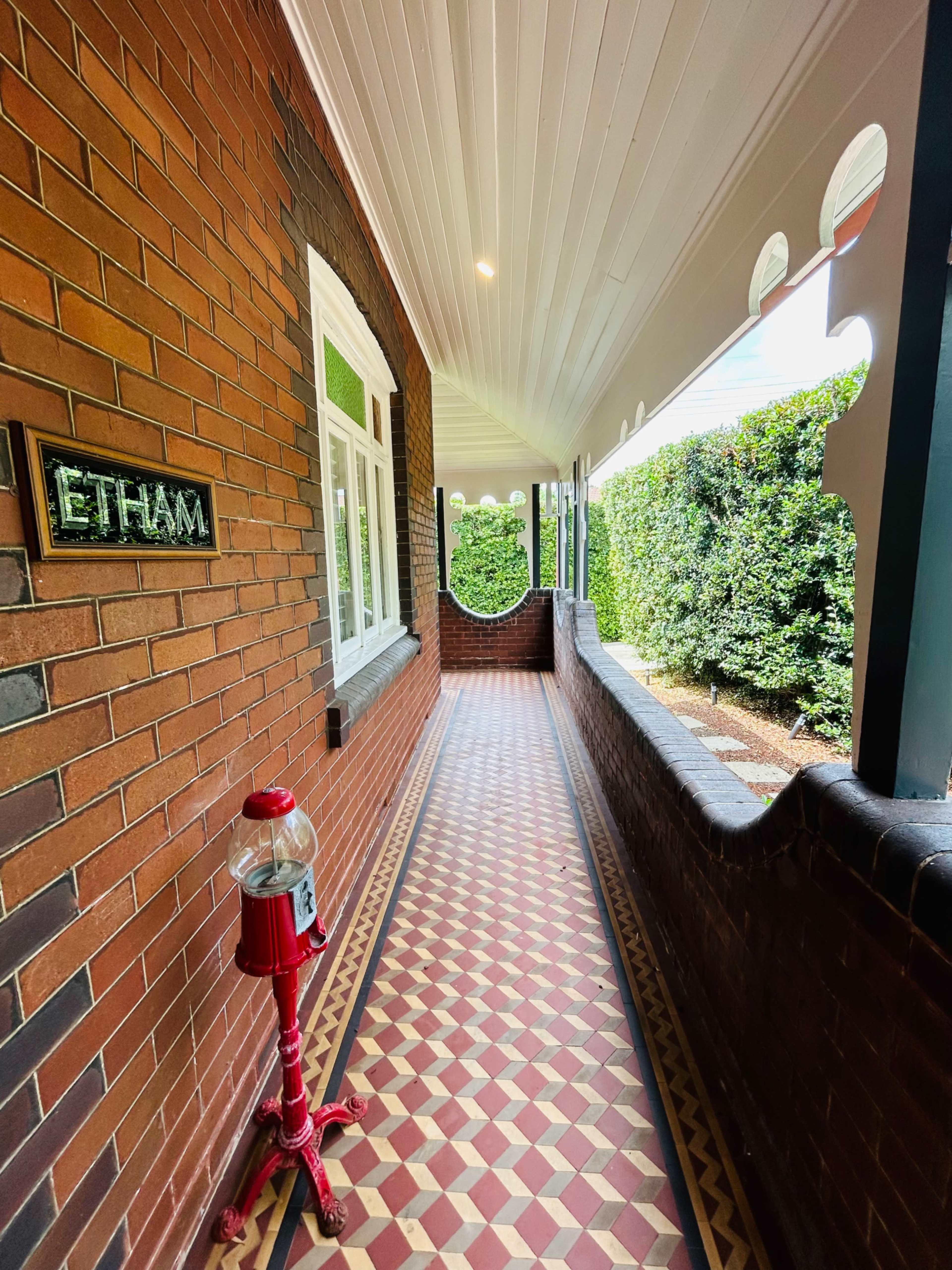 A narrow corridor with a patterned tile floor, bordered by brick walls and topped with a decorative overhang, features a vintage candy dispenser on the left and green hedges visible at the end.