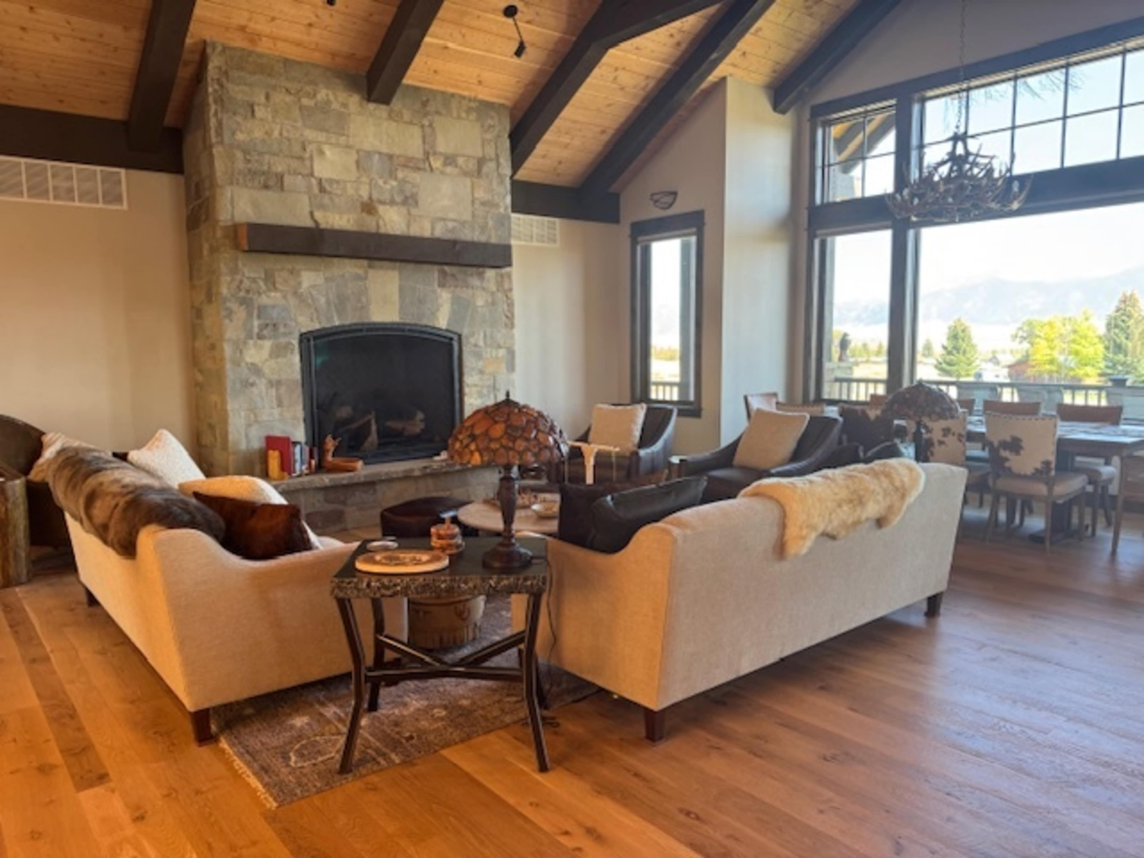 The image shows a spacious living room featuring two sofas, a stone fireplace, large windows with a view of mountains, and a dining area with chairs in the background.