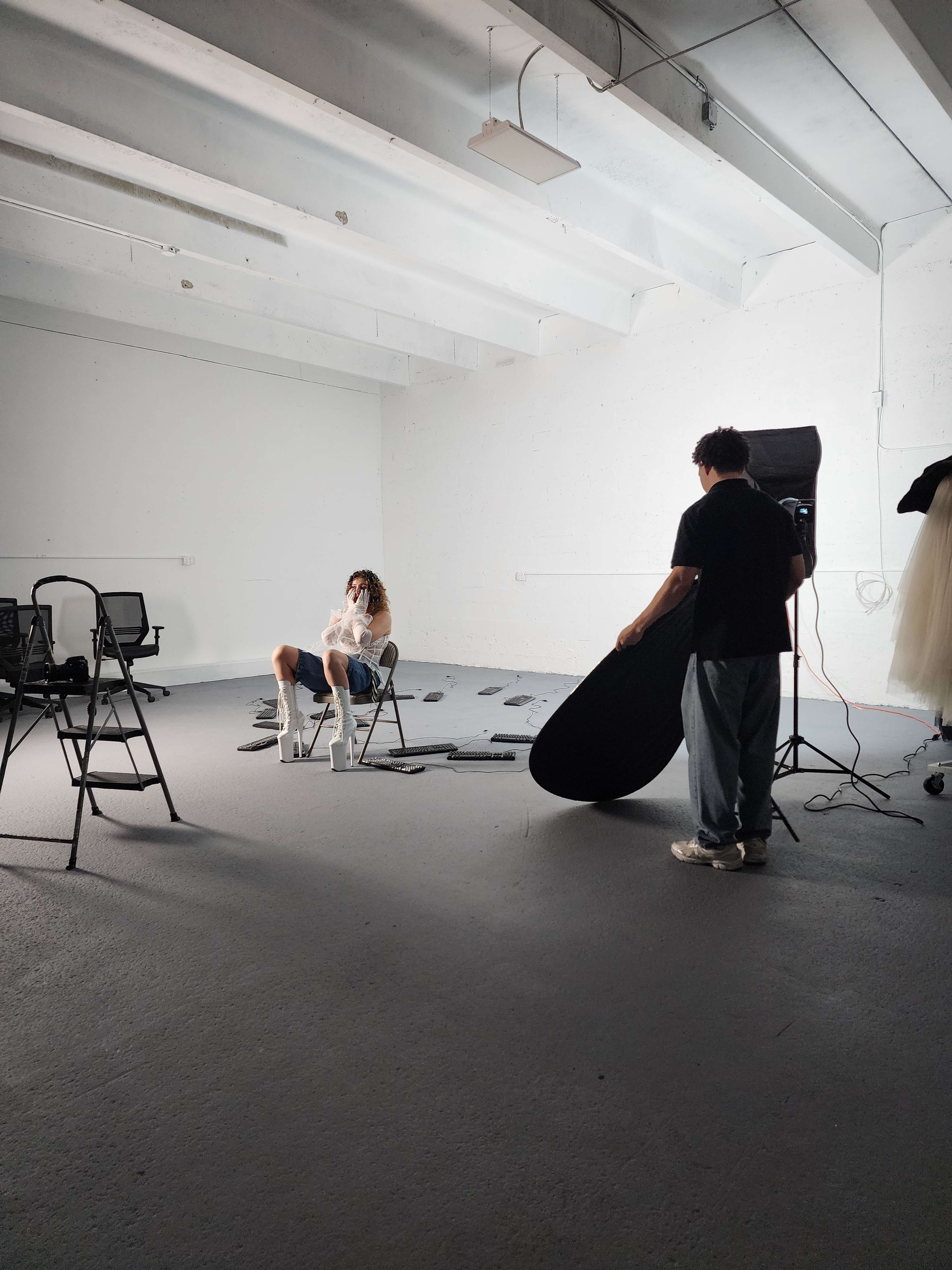 A photographer adjusts a reflector while a model sits on a chair in a sparsely furnished studio with bright lighting.