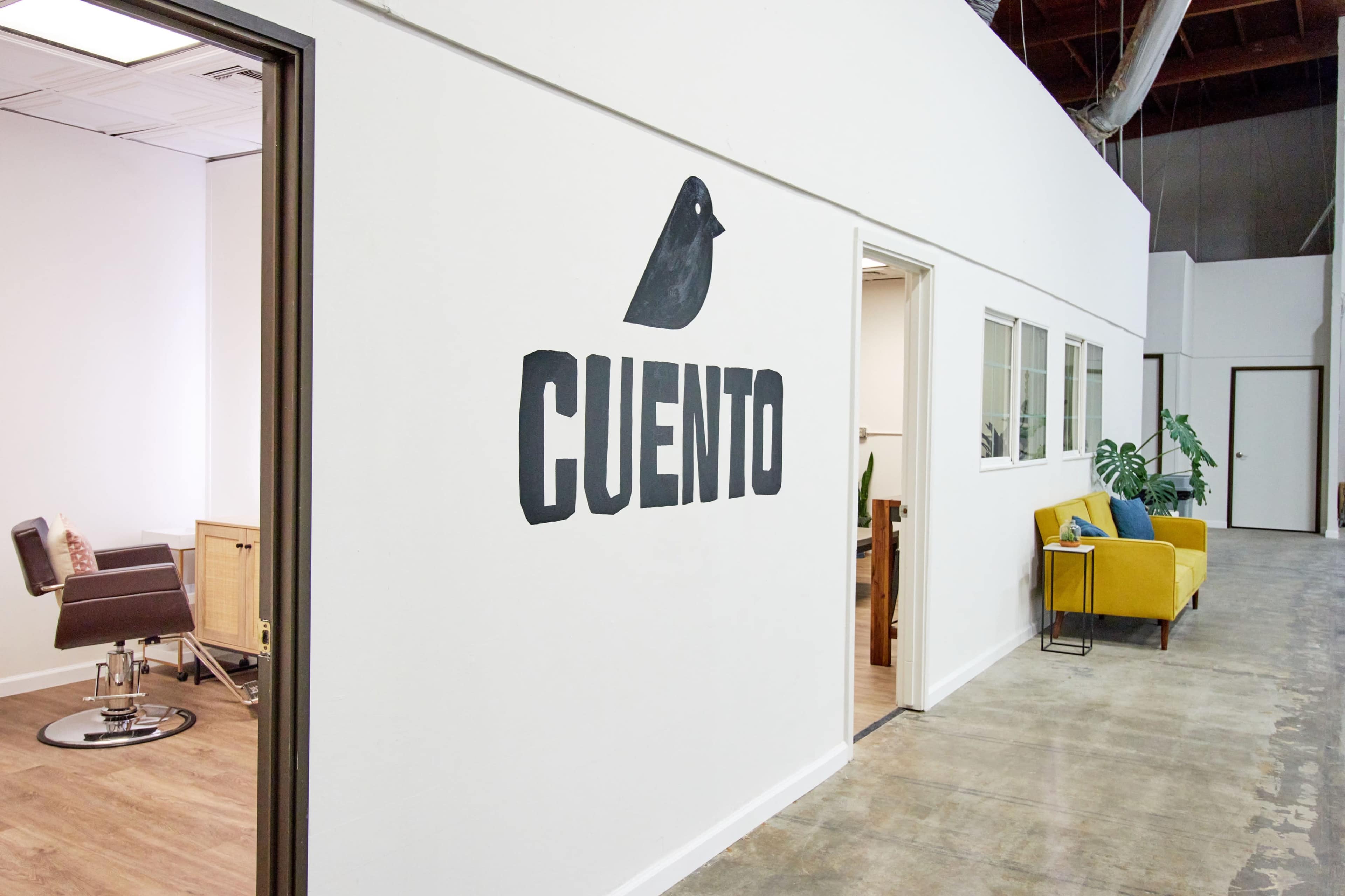 A modern office hallway with a large sign reading "CUENTO" on the wall, an office door on the left, and a yellow couch visible in the background.