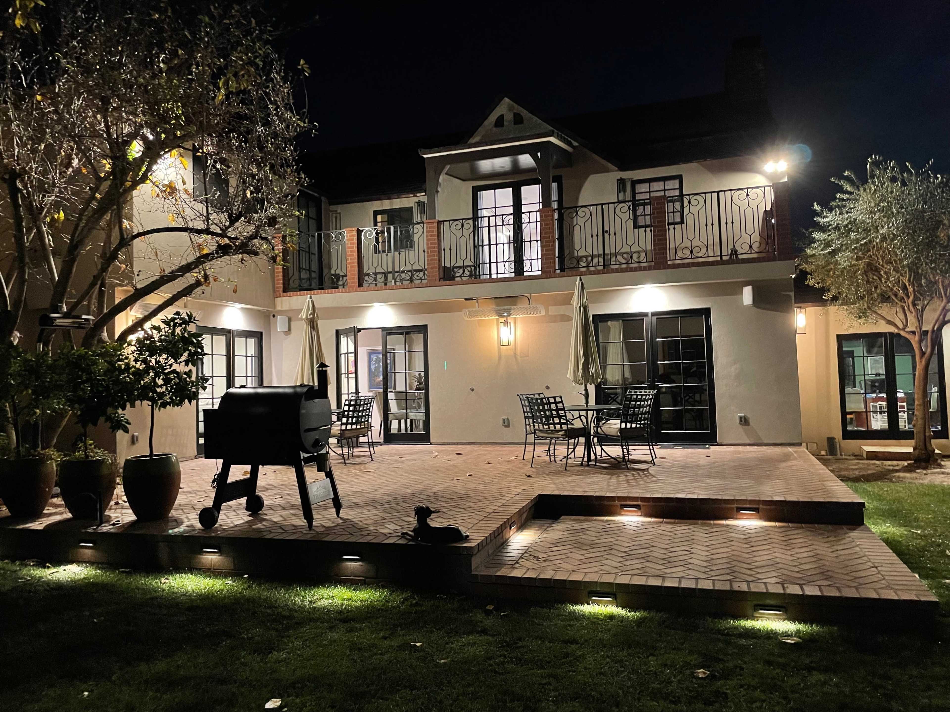 A well-lit backyard patio featuring a grill, outdoor seating, and surrounded by landscaped greenery at night.