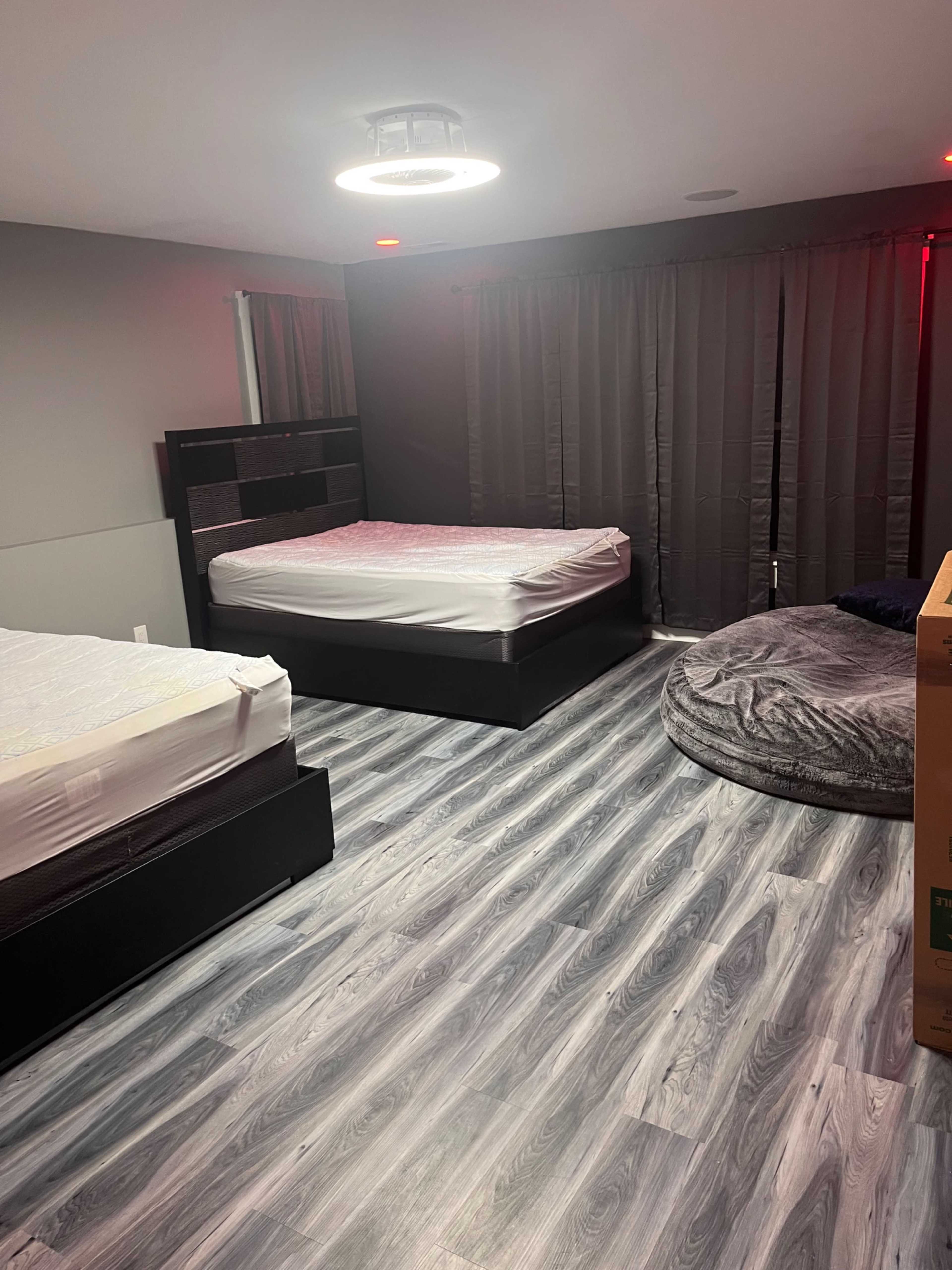 A room with two unmade beds, a round gray beanbag chair, and dark curtains covering the windows.
