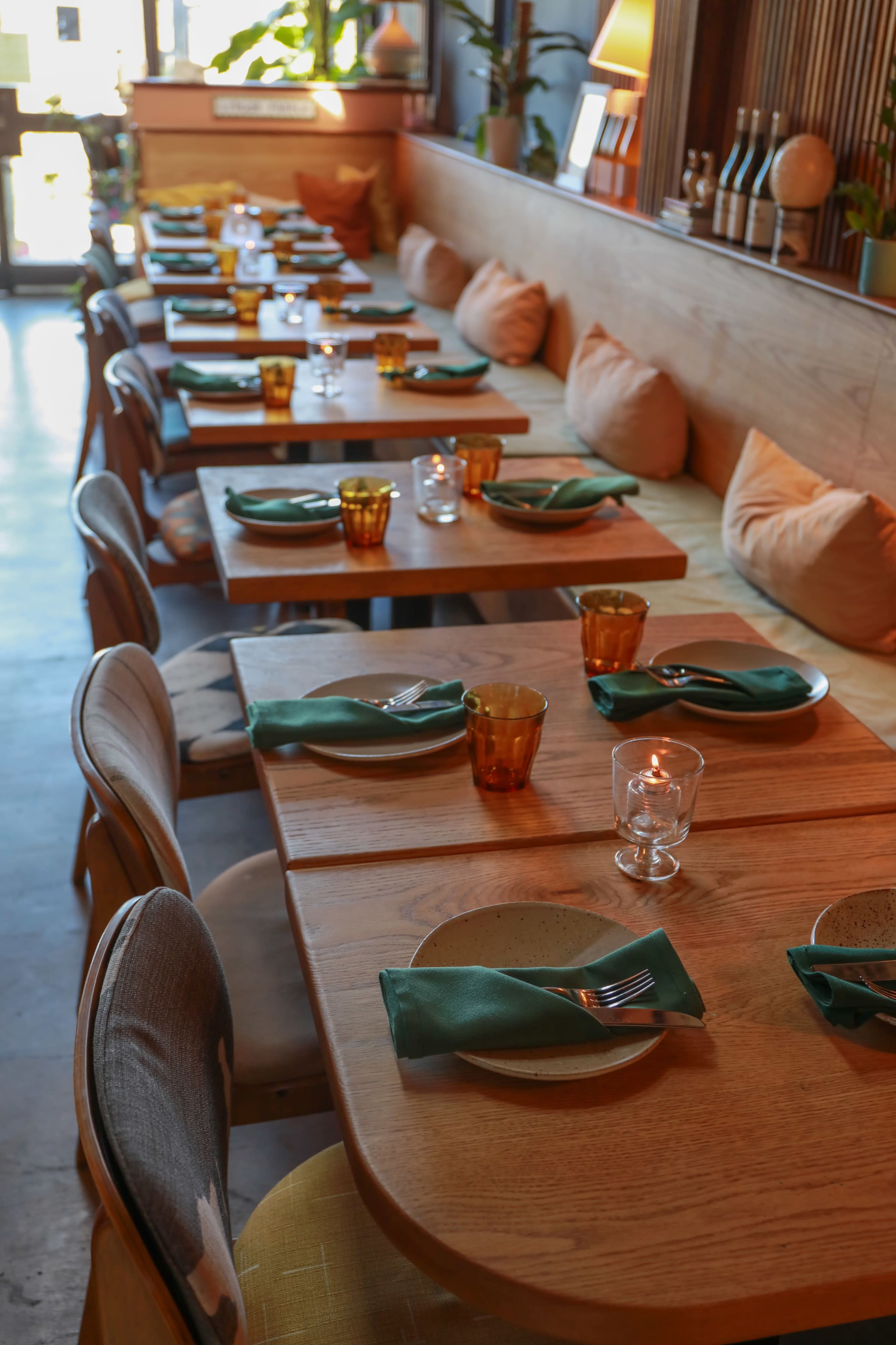 The image shows a restaurant interior with several wooden tables set with green napkins, cutlery, and amber-colored glasses, flanked by cushioned benches.