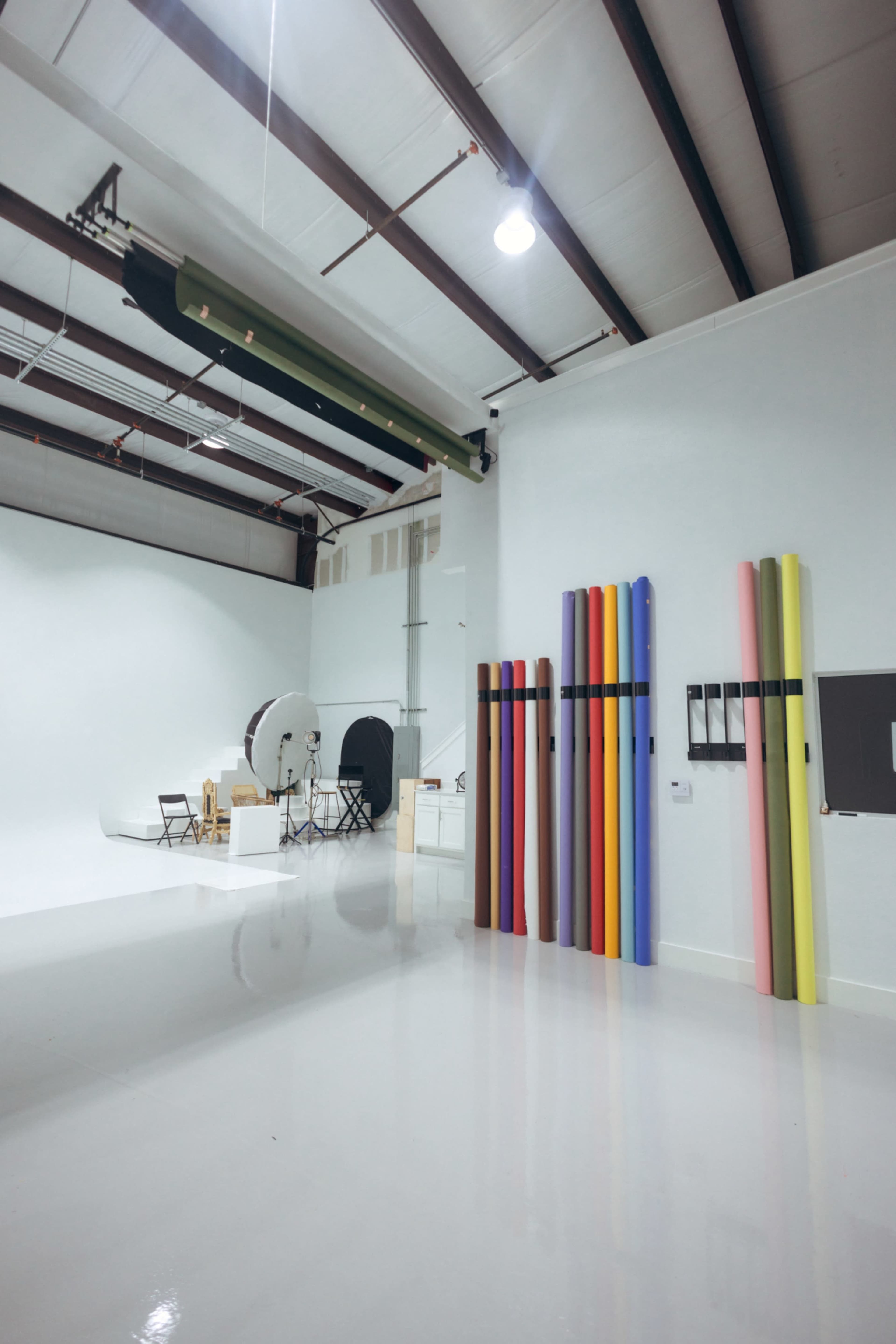 The image shows a photography studio with a white backdrop, various lighting equipment, and colorful backdrops arranged vertically against a wall.
