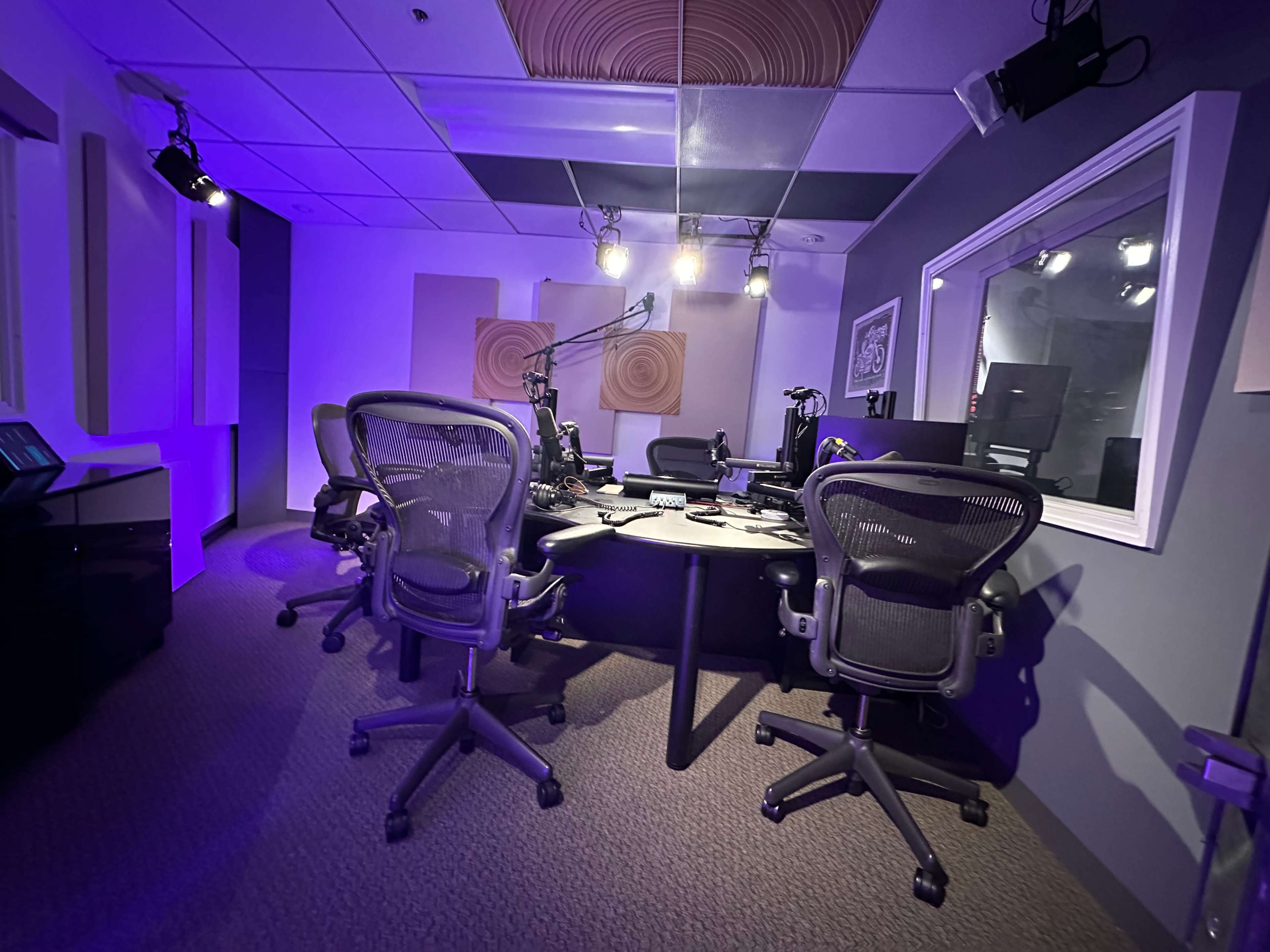 A radio studio is equipped with multiple chairs, microphones, and sound equipment, with purple lighting illuminating the space.