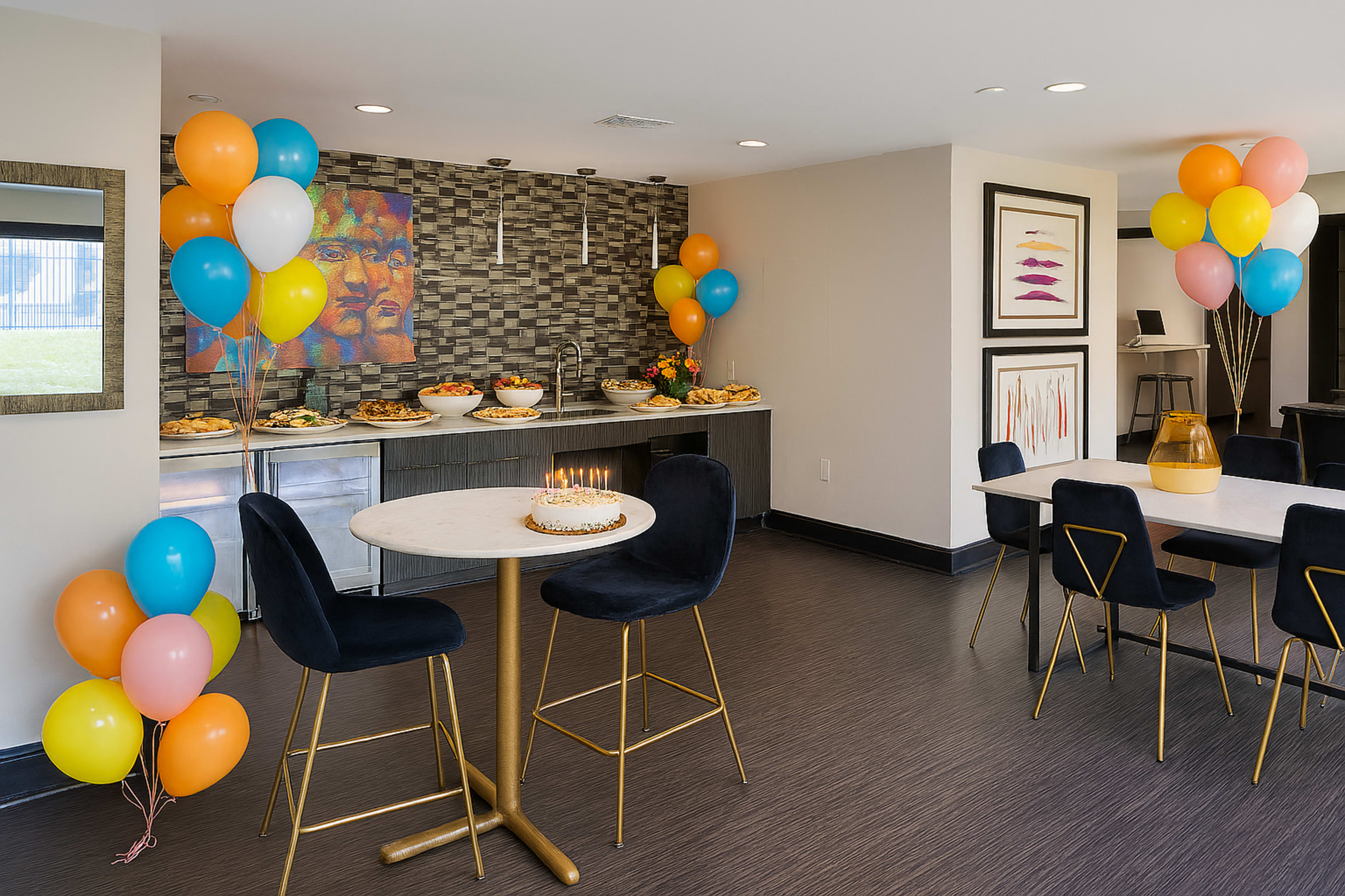 A decorated event space features a table with food, a small cake, and colorful balloons lining the walls, along with seating arrangements.