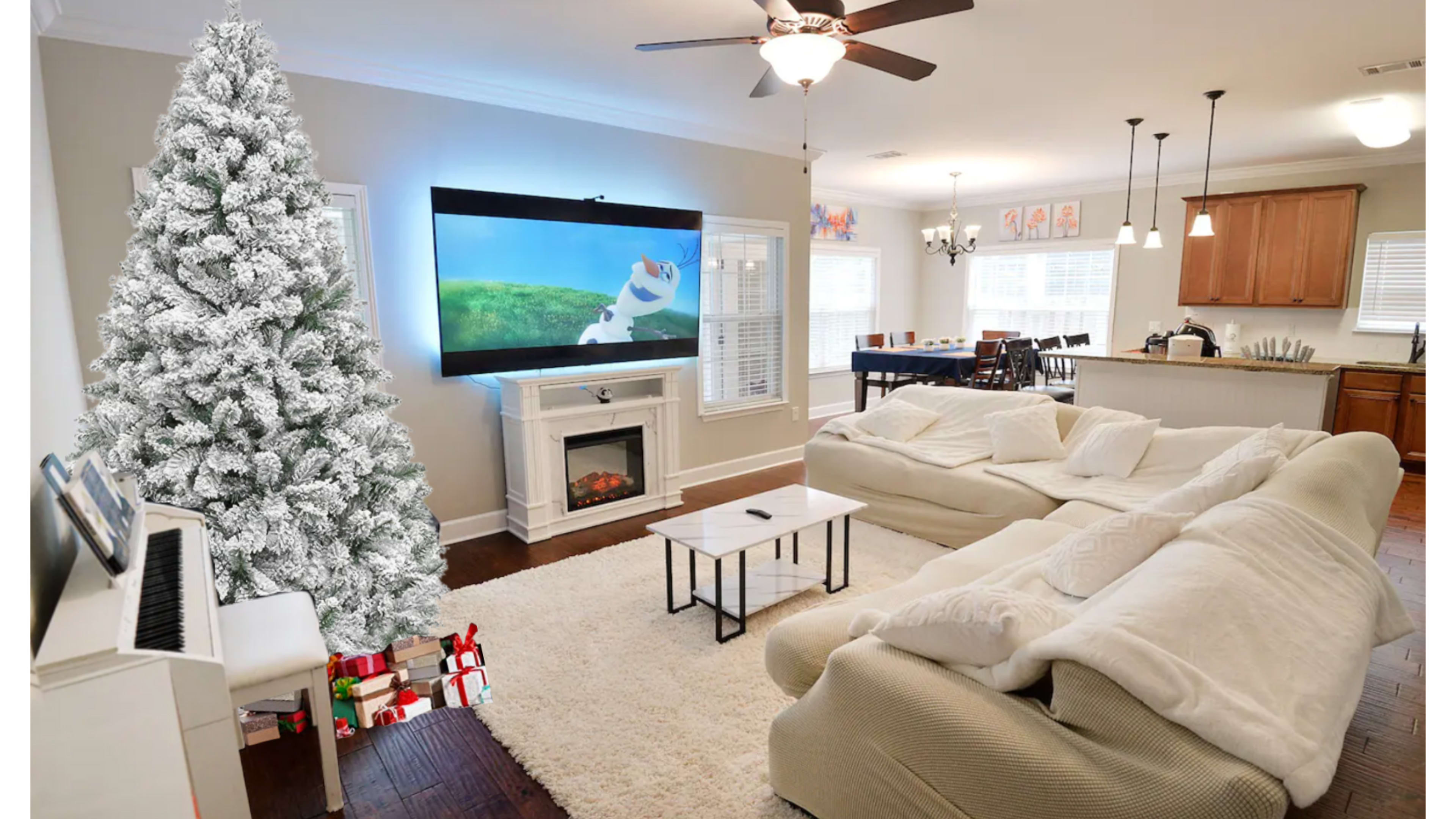 The living room features a large white artificial Christmas tree next to a sectional sofa, a coffee table, a fireplace with a TV mounted above it, and a dining area in the background.