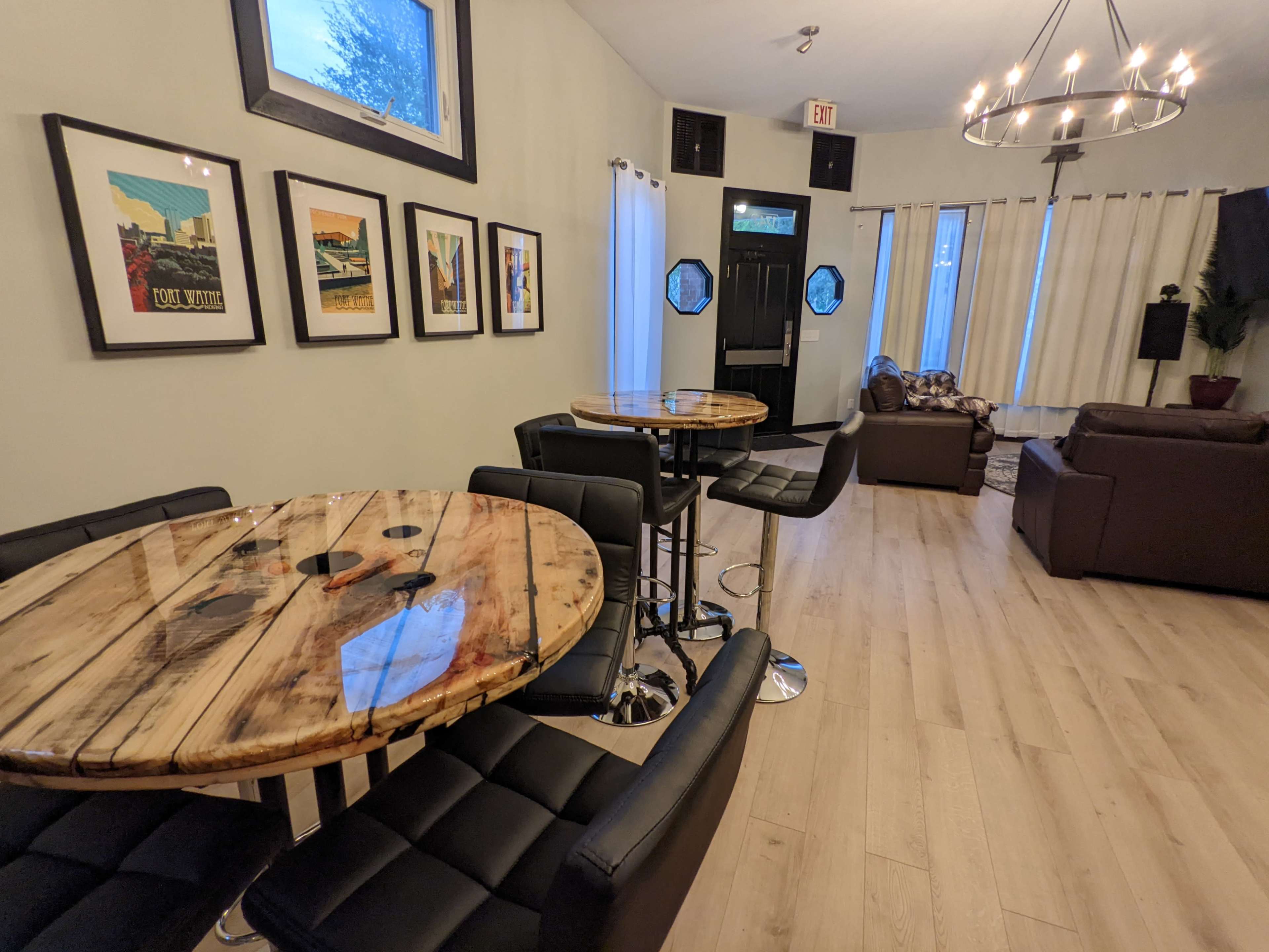 A modern interior space with two wooden tables surrounded by black chairs, framed artwork on the walls, and a seating area with a brown couch and curtains.
