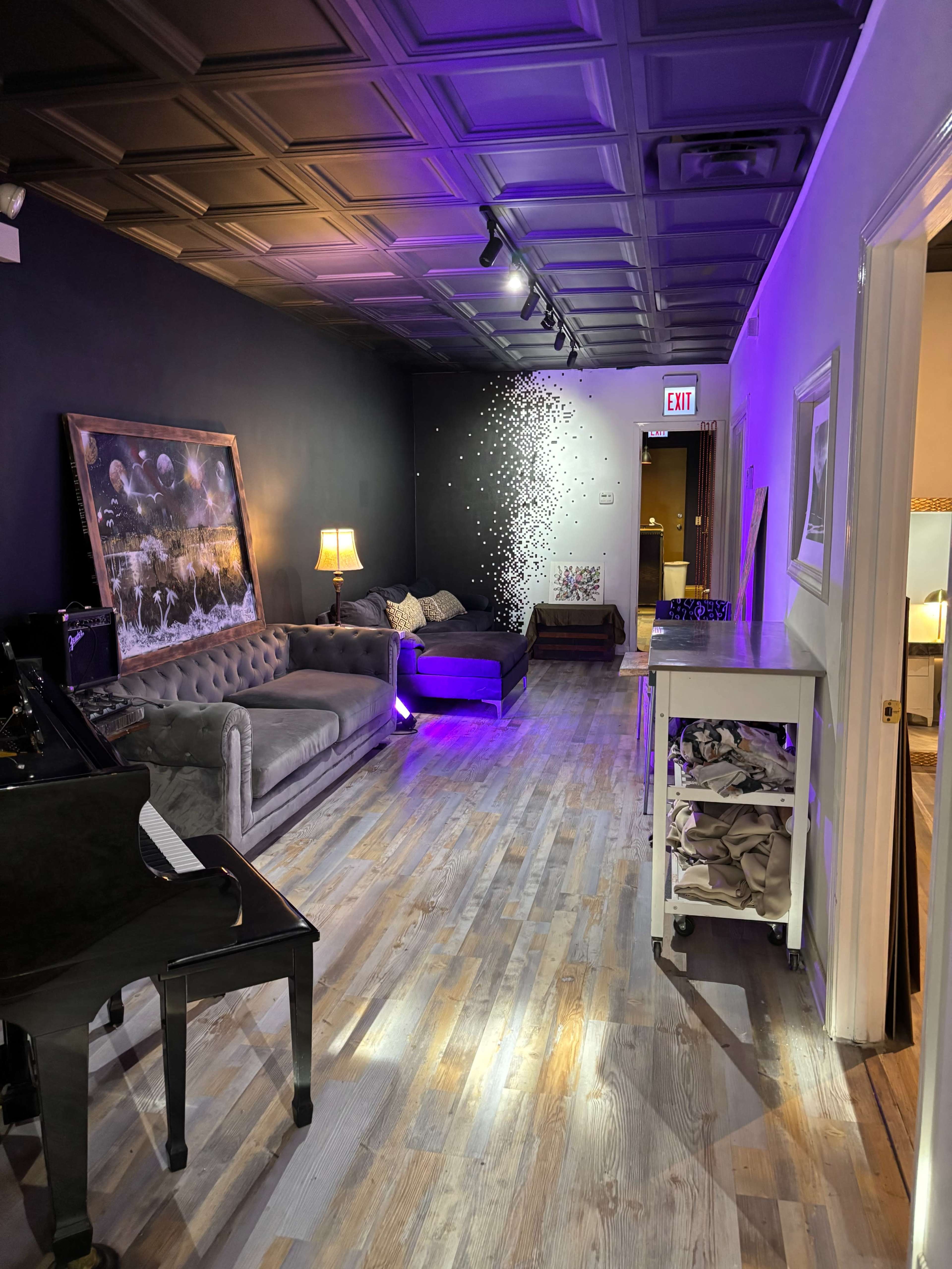 The image shows a narrow room with wooden flooring, featuring two sofas, a coffee table, a lamp, and a piano in one corner, with a wall adorned with artwork and a splash of decorative light effects.