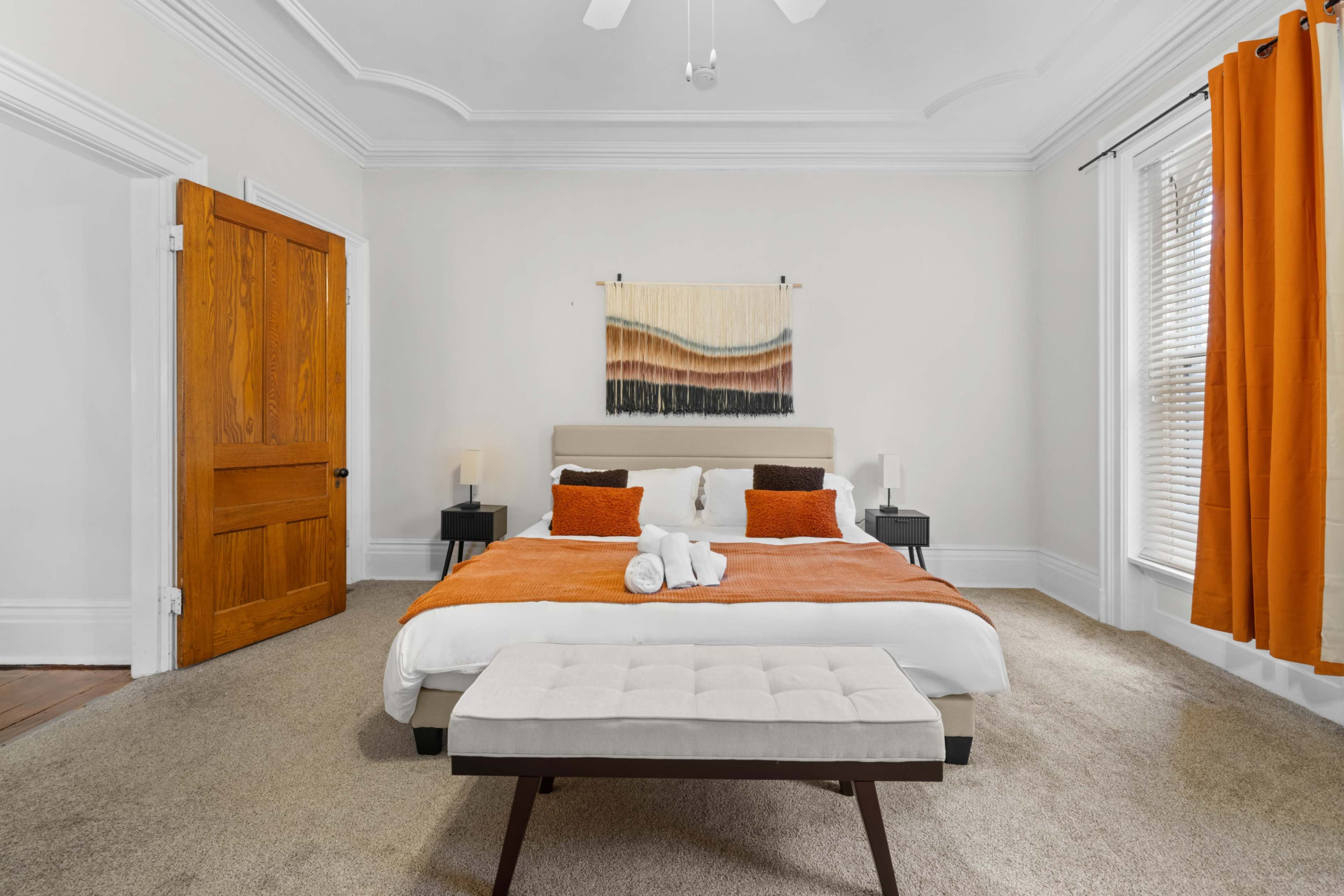 The image shows a neatly arranged bedroom featuring a bed with orange and white bedding, a bench at the foot of the bed, and large windows with orange curtains.