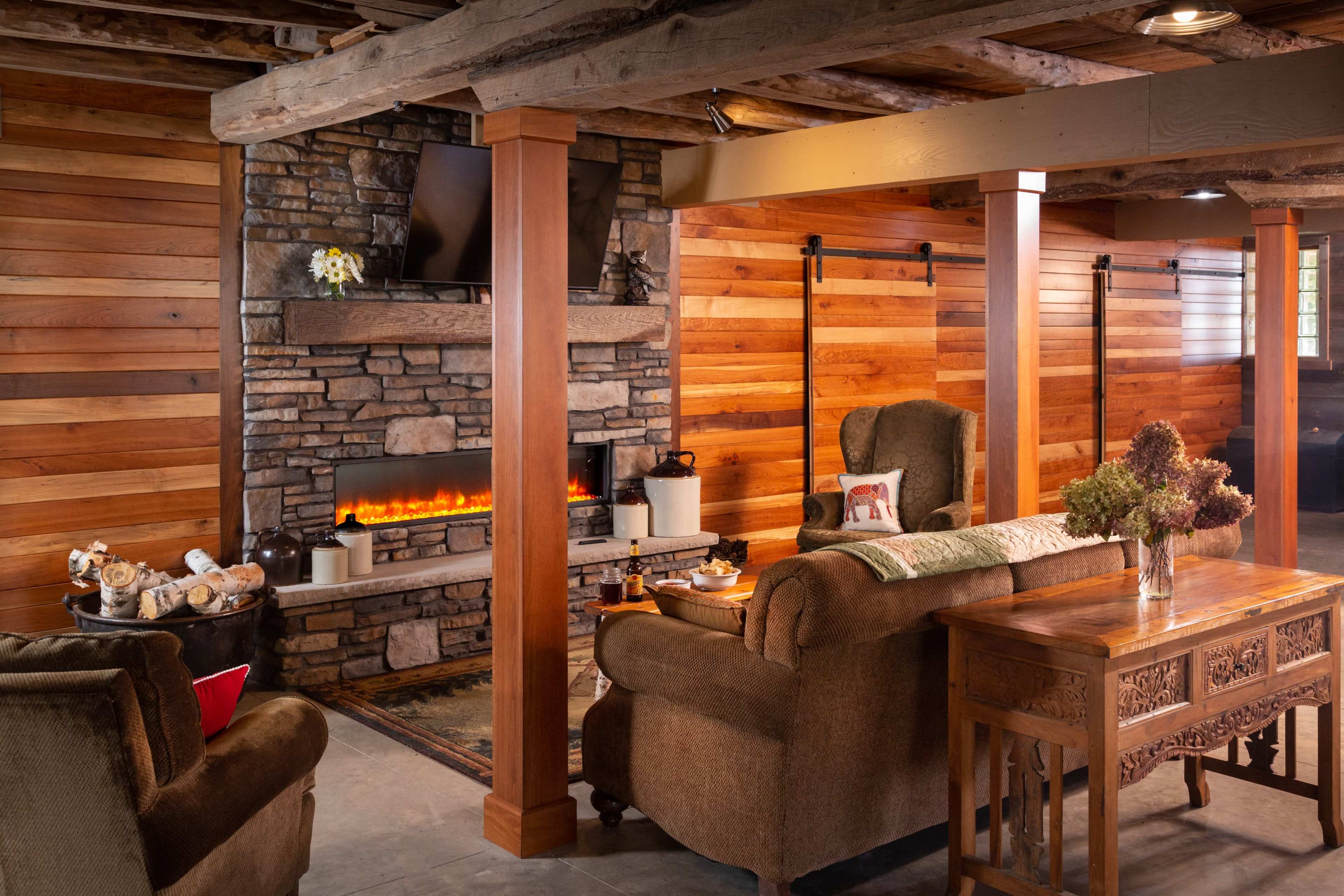 The image depicts a cozy living room featuring a stone fireplace, wooden walls, and comfortable seating arrangements.