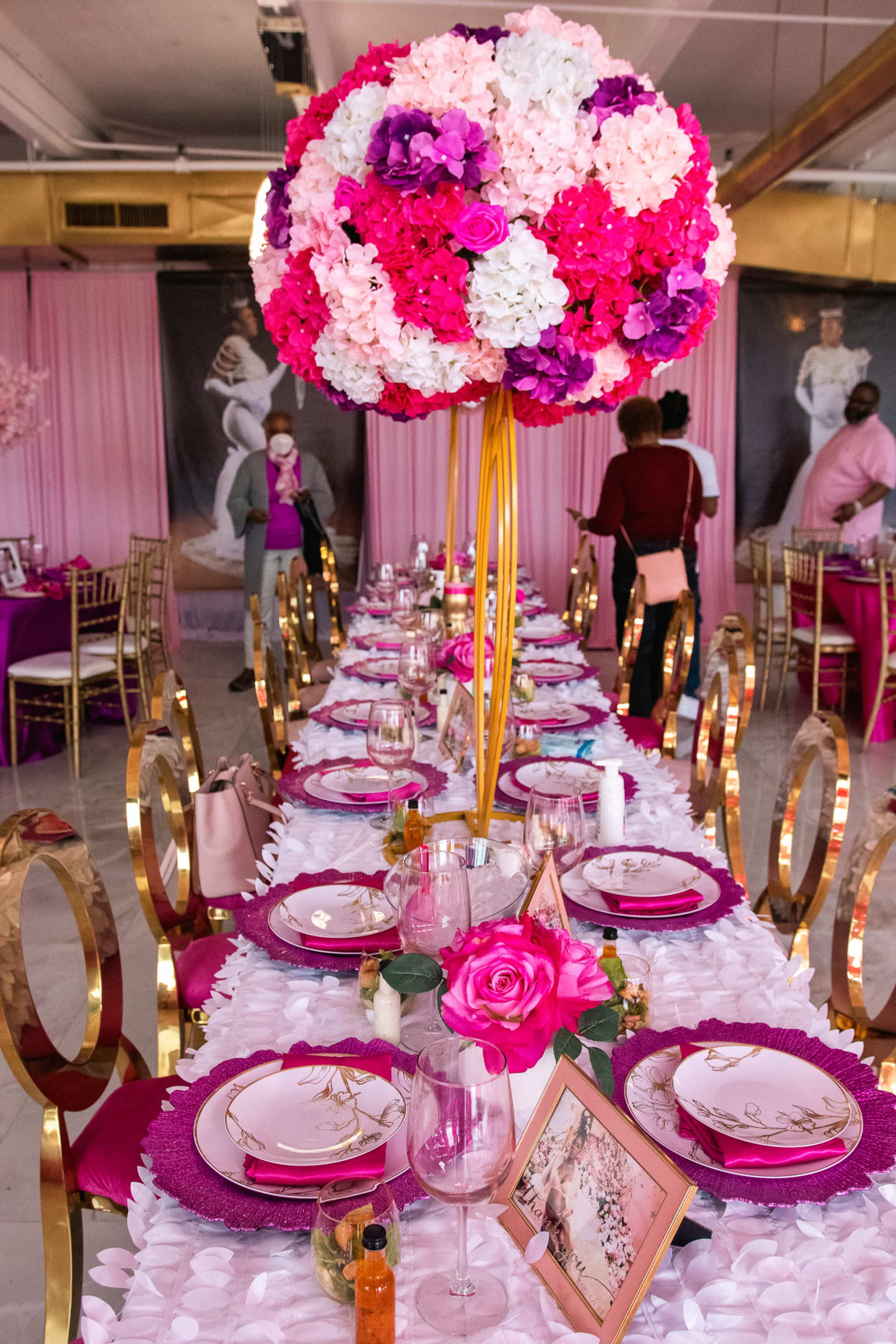 A decorated dining table set with pink and purple floral arrangements, elegant tableware, and a backdrop featuring fashion photos.