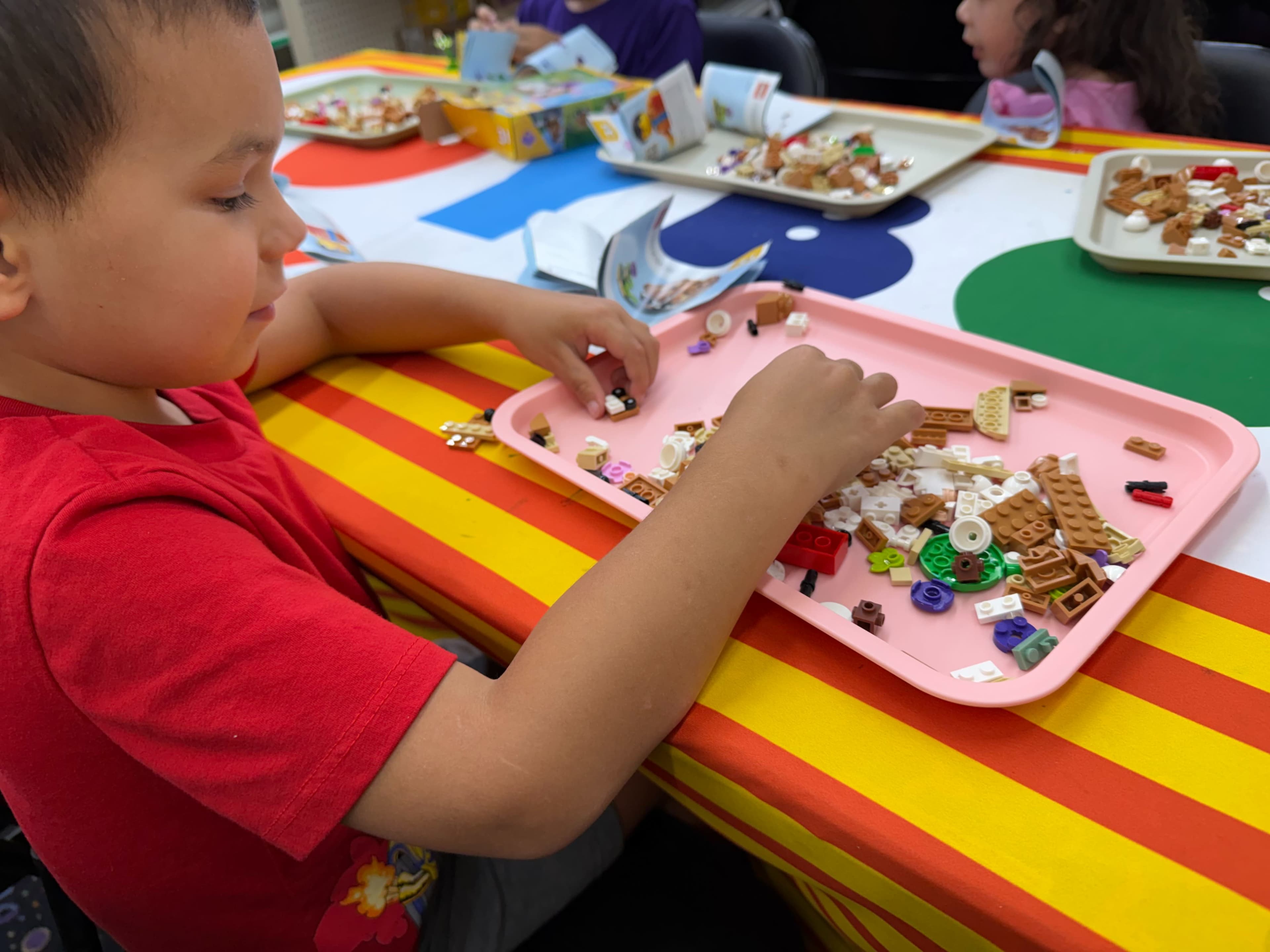 A young child is assembling colorful building blocks on a tray while seated at a table decorated with a vibrant striped tablecloth.