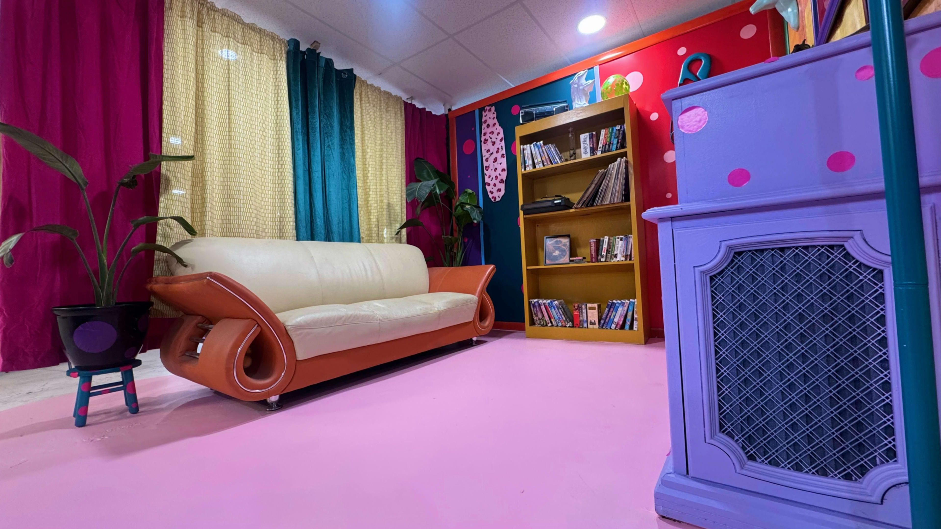 The image shows a vibrant interior with a brown and cream sofa, a wooden bookshelf filled with books and DVDs, colorful walls, and a pink floor.