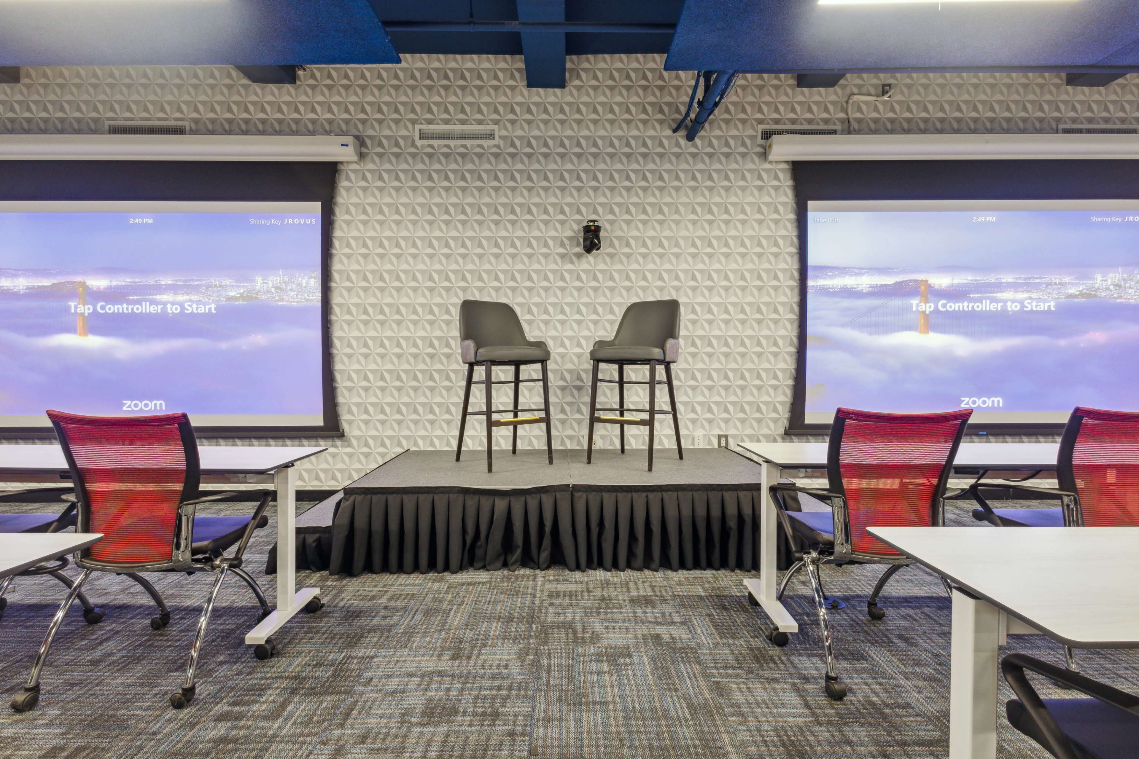 A presentation area with two high chairs on a stage, flanked by large screens displaying the Zoom logo and instructions to "Tap Controller to Start."