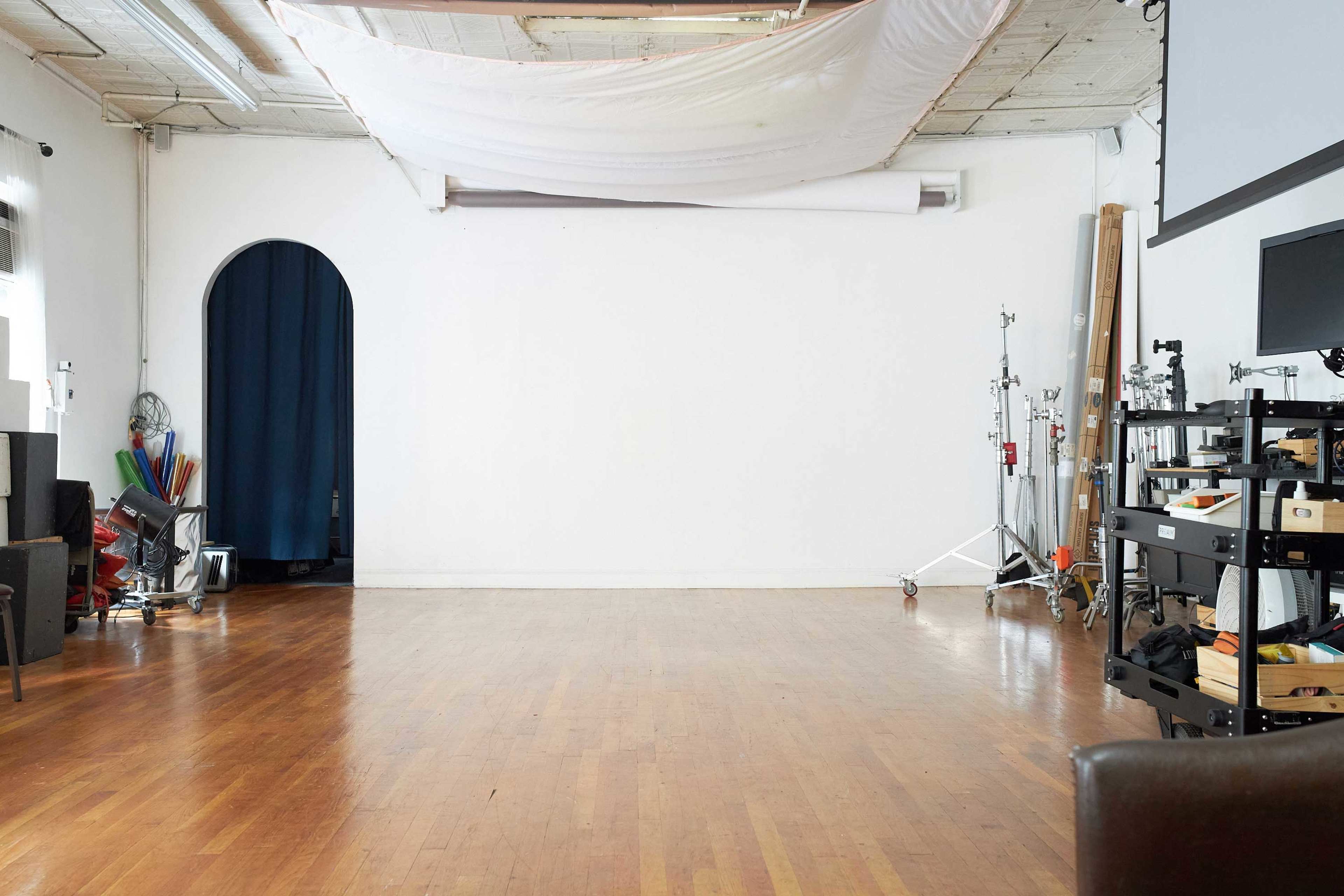 The image shows a spacious, empty studio with wooden flooring, a white wall, and various equipment and props arranged along the sides.