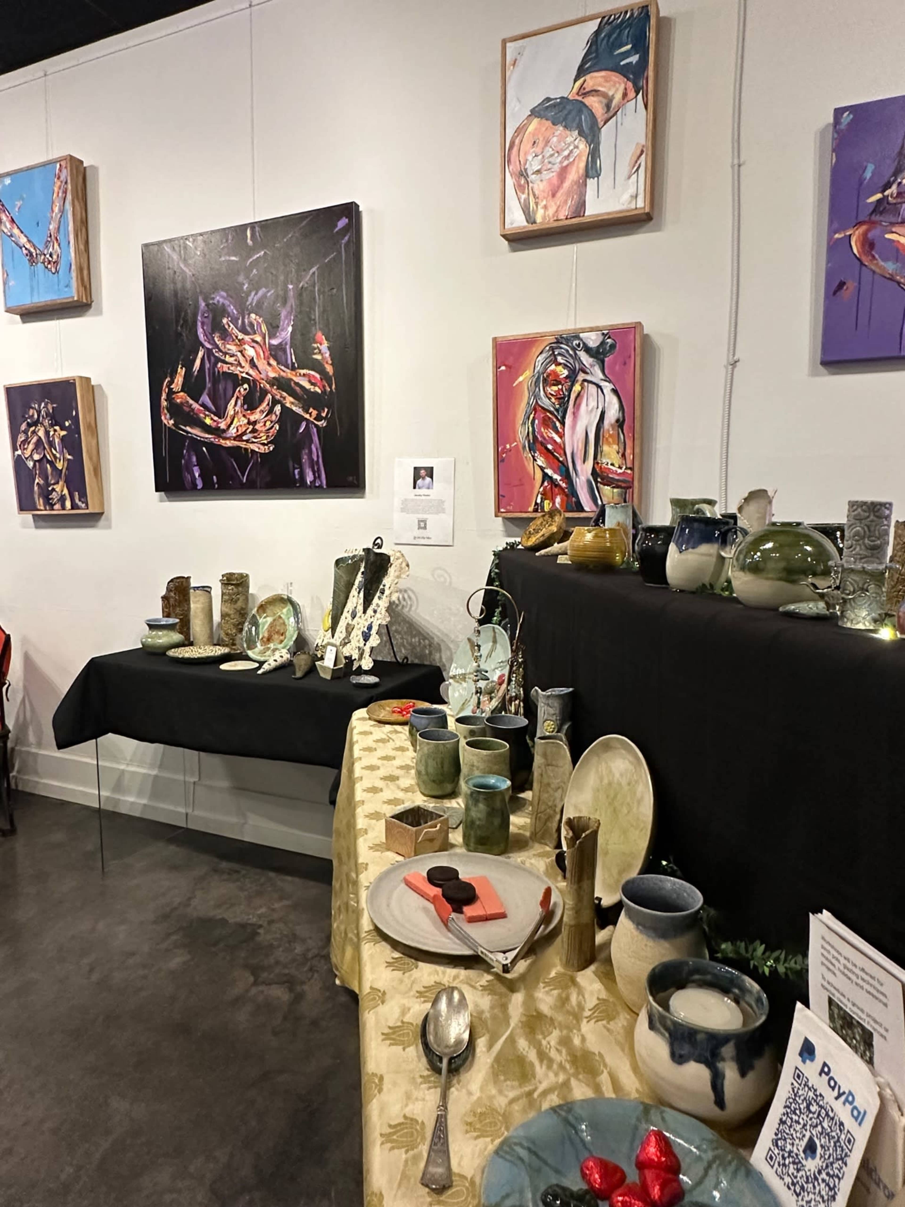 A display of ceramic art pieces is set on a table beneath colorful paintings in a gallery.