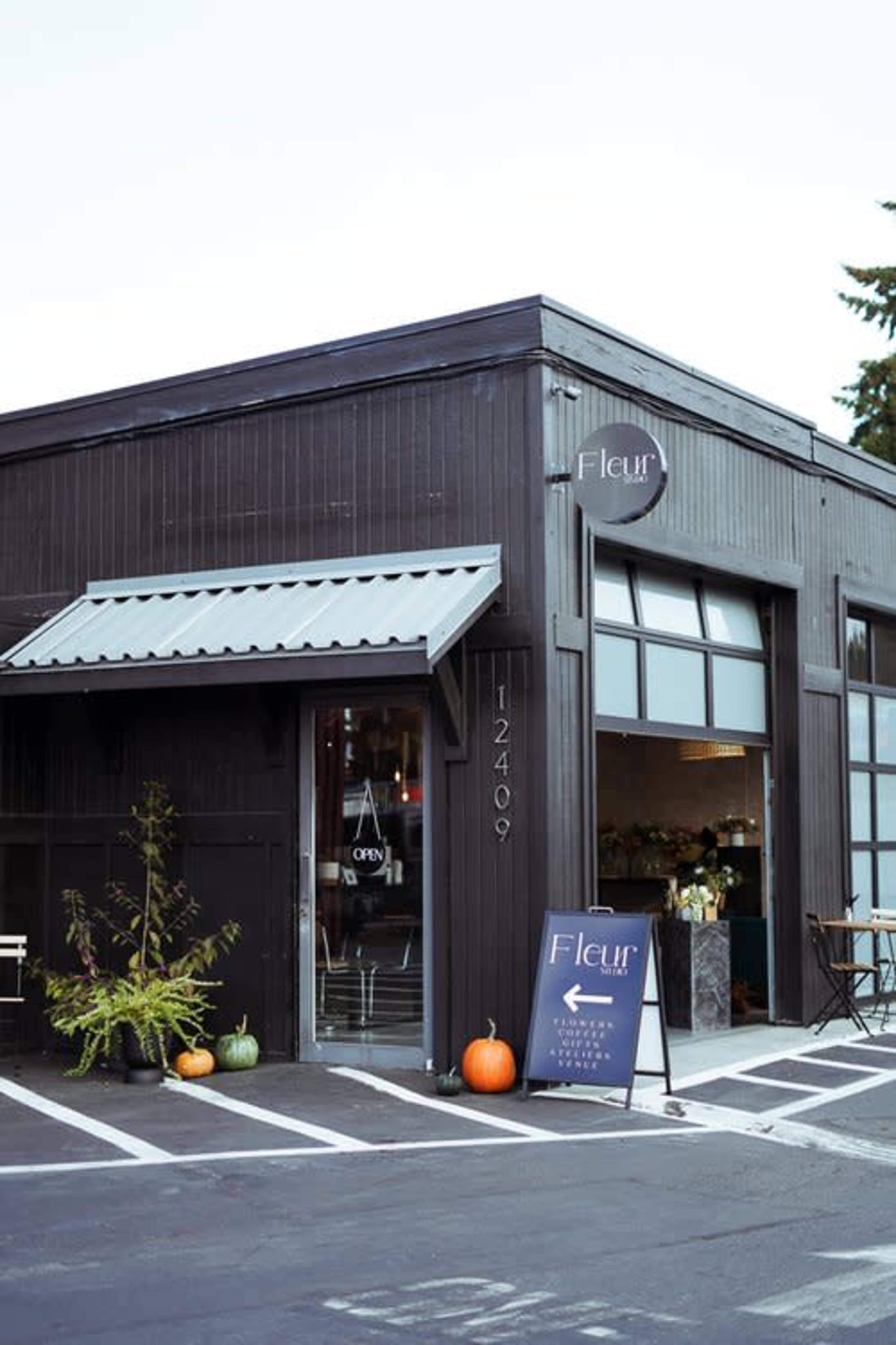 A black wooden flower shop named "Fleur" features an open sign, pumpkins outside, and a directional sign on the side of the building.