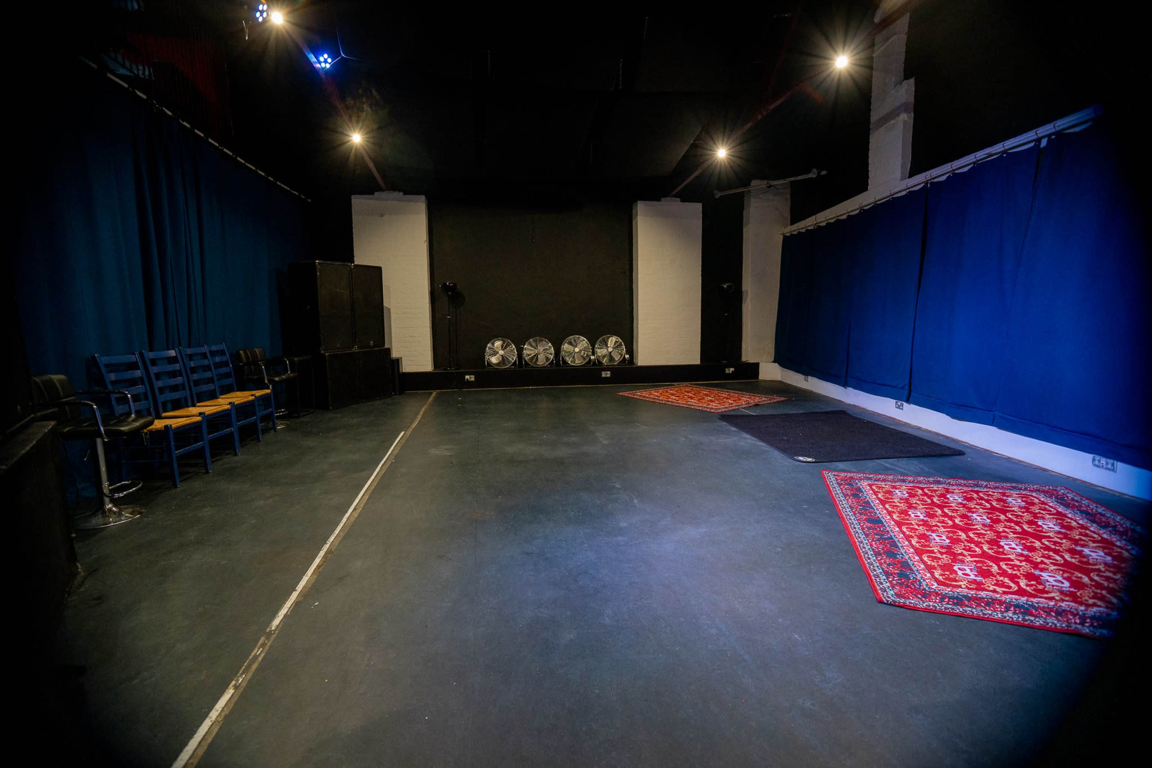 The image shows an empty performance space with blue curtains, chairs along one side, and three large fans arranged near the back wall.