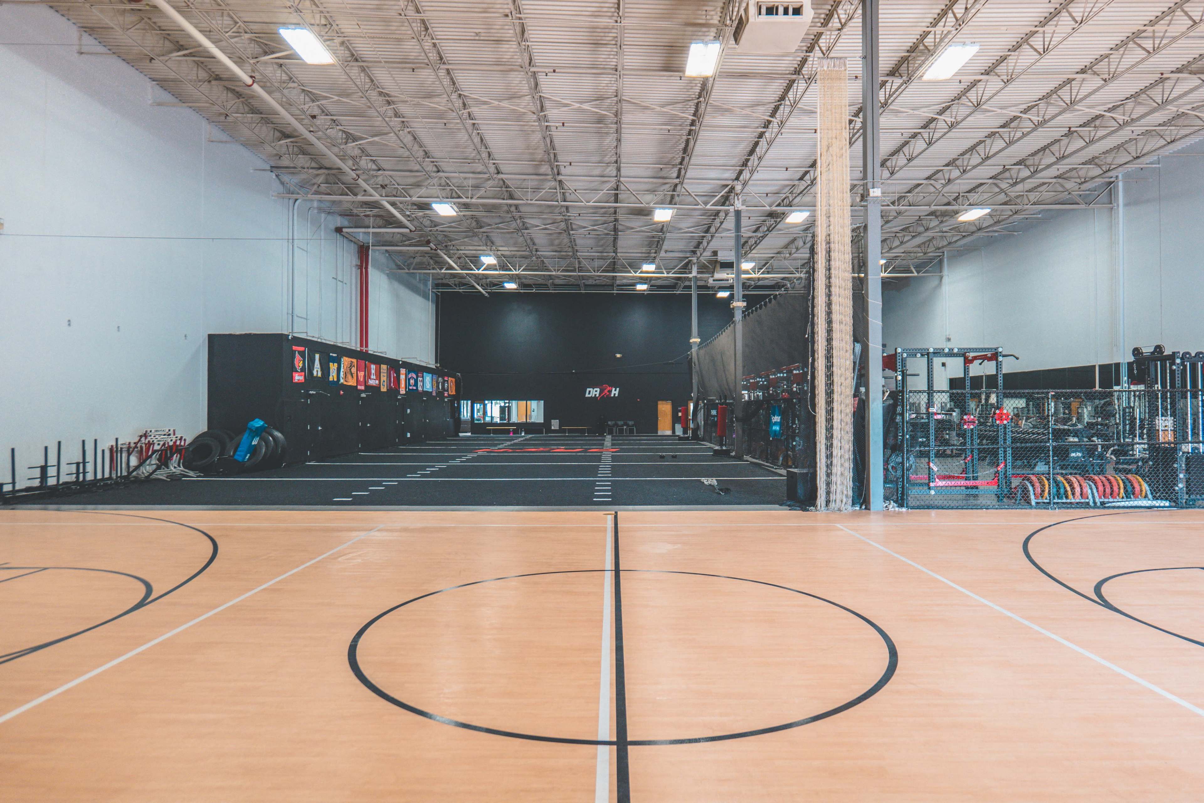 A spacious indoor gymnasium with a wooden floor, marked for basketball, and workout equipment visible in the background.