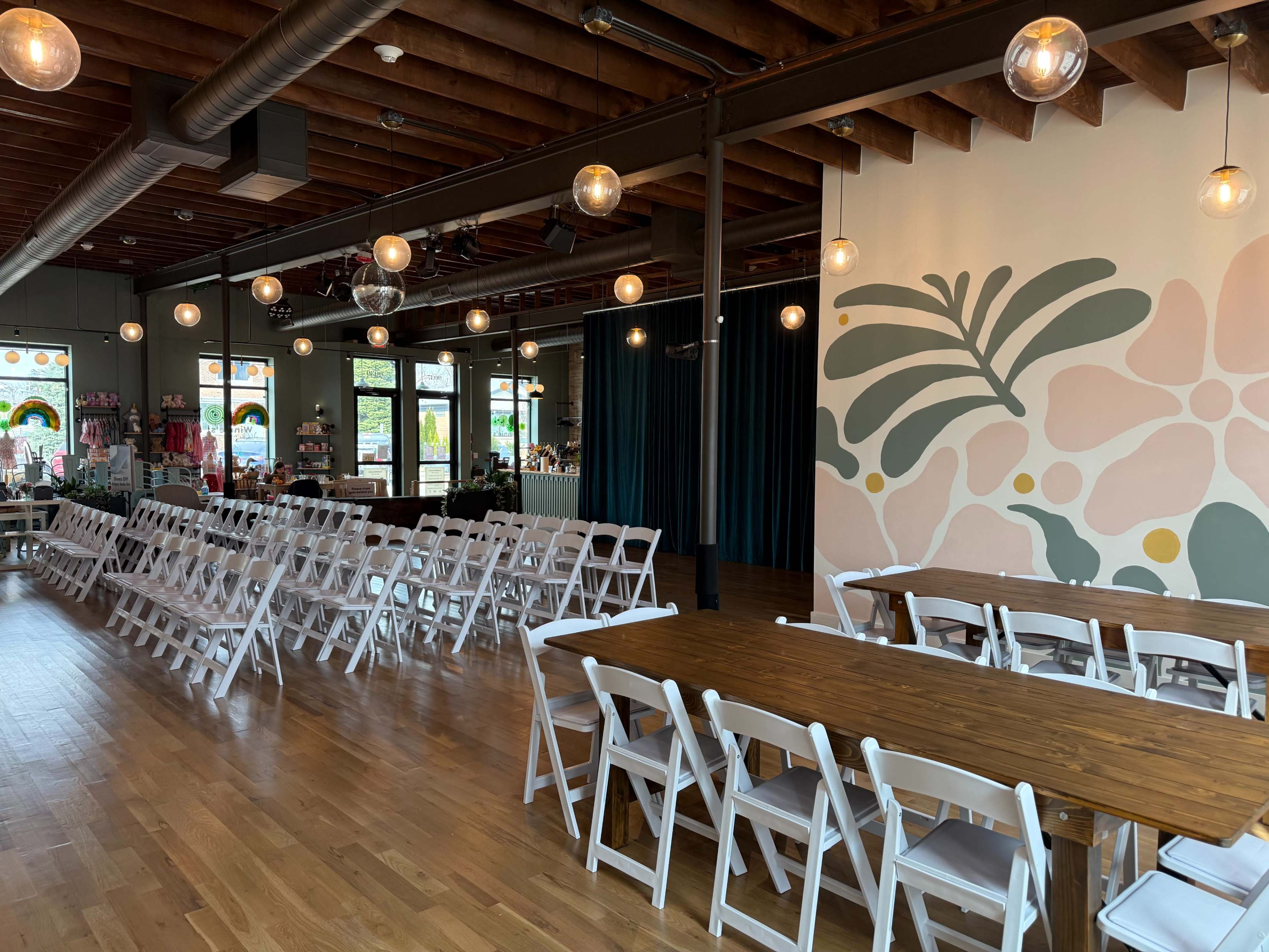 The image shows an interior space set up for an event, featuring rows of white folding chairs and a long wooden table, with decorative light fixtures and a colorful mural on one wall.