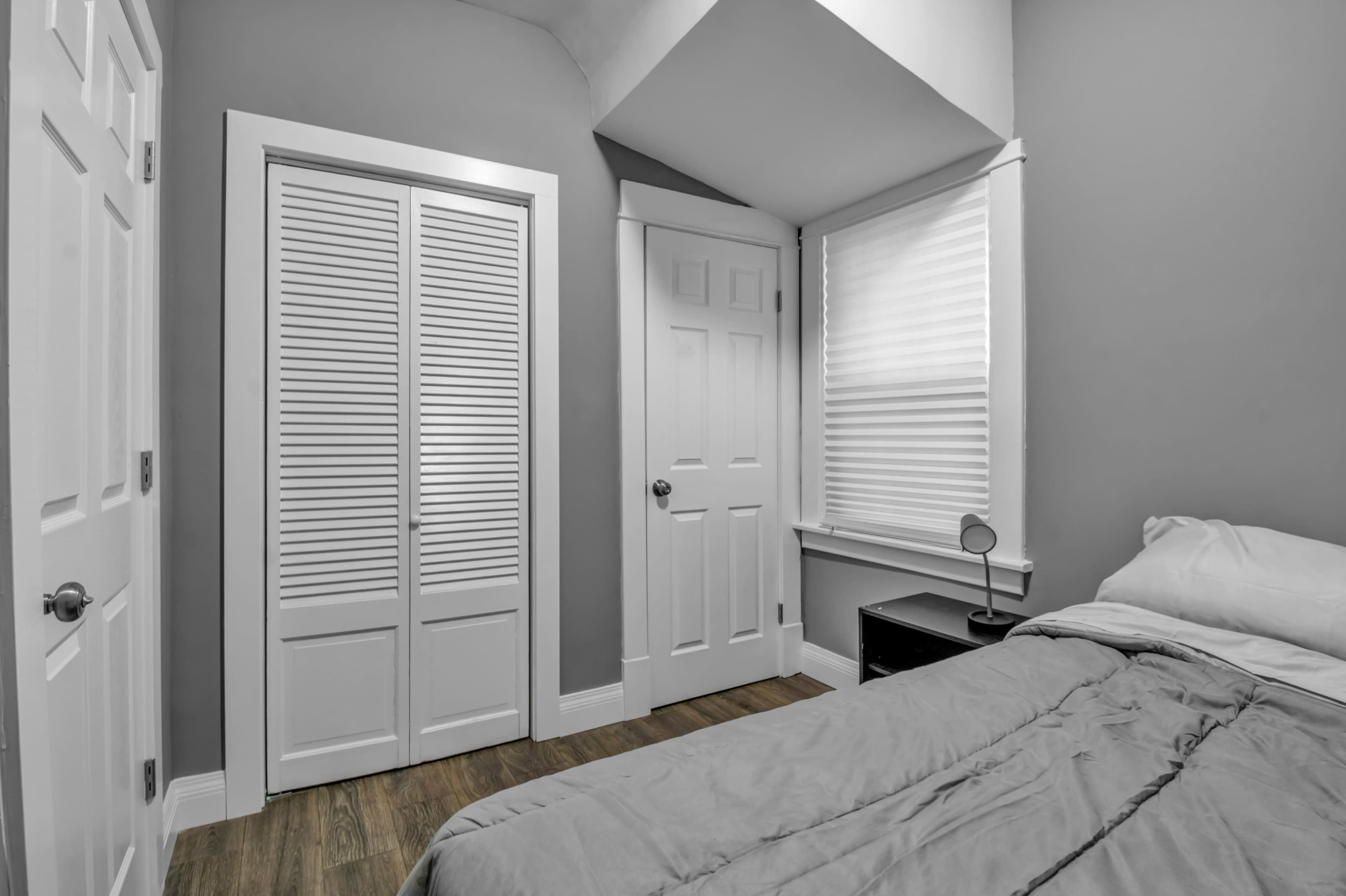 A small bedroom features a bed, a nightstand, and a window with blinds, alongside closet and door entrances.