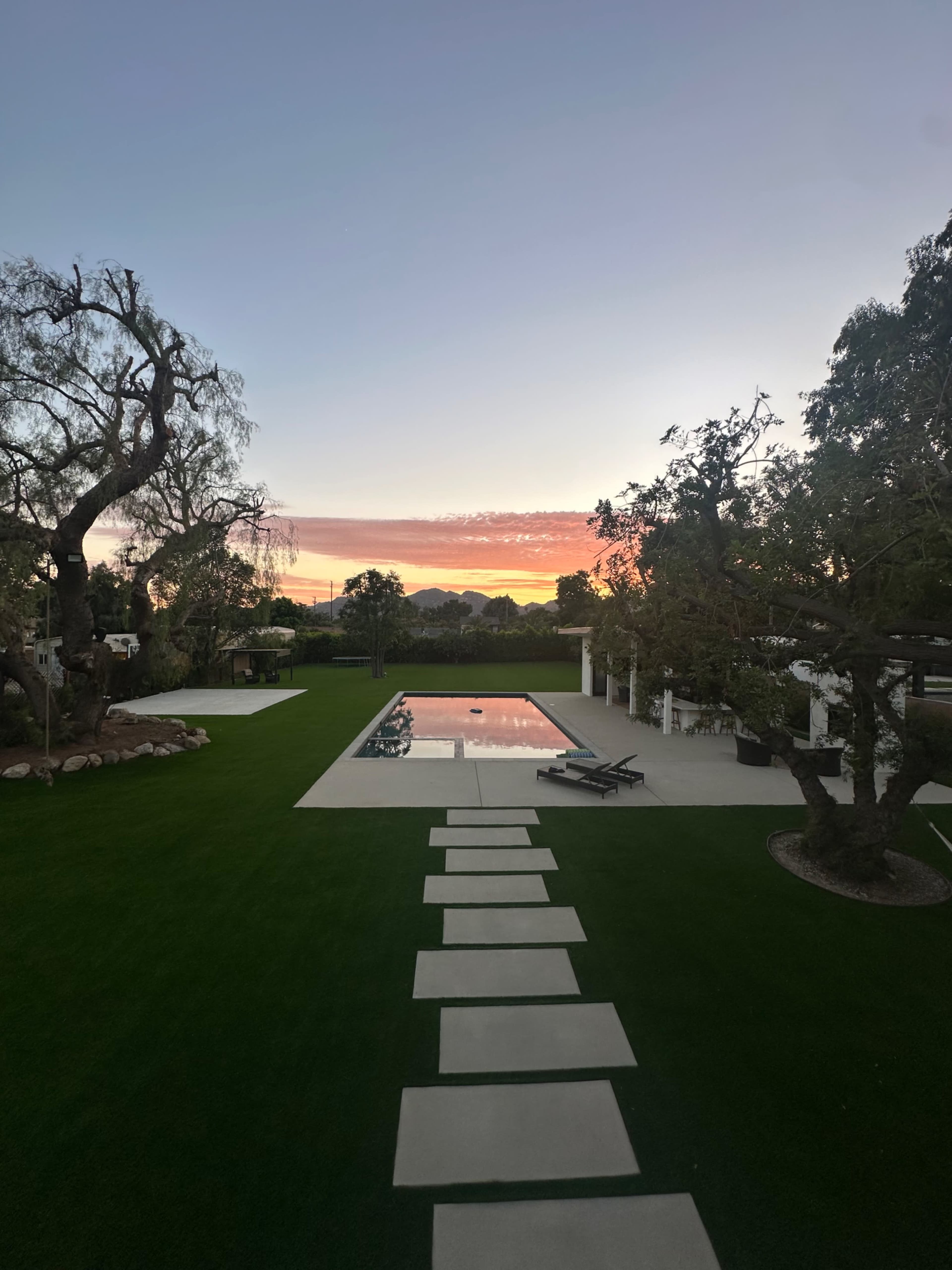 The image shows a landscaped backyard with a swimming pool reflecting the sunset and stone pathways leading across a grassy area.