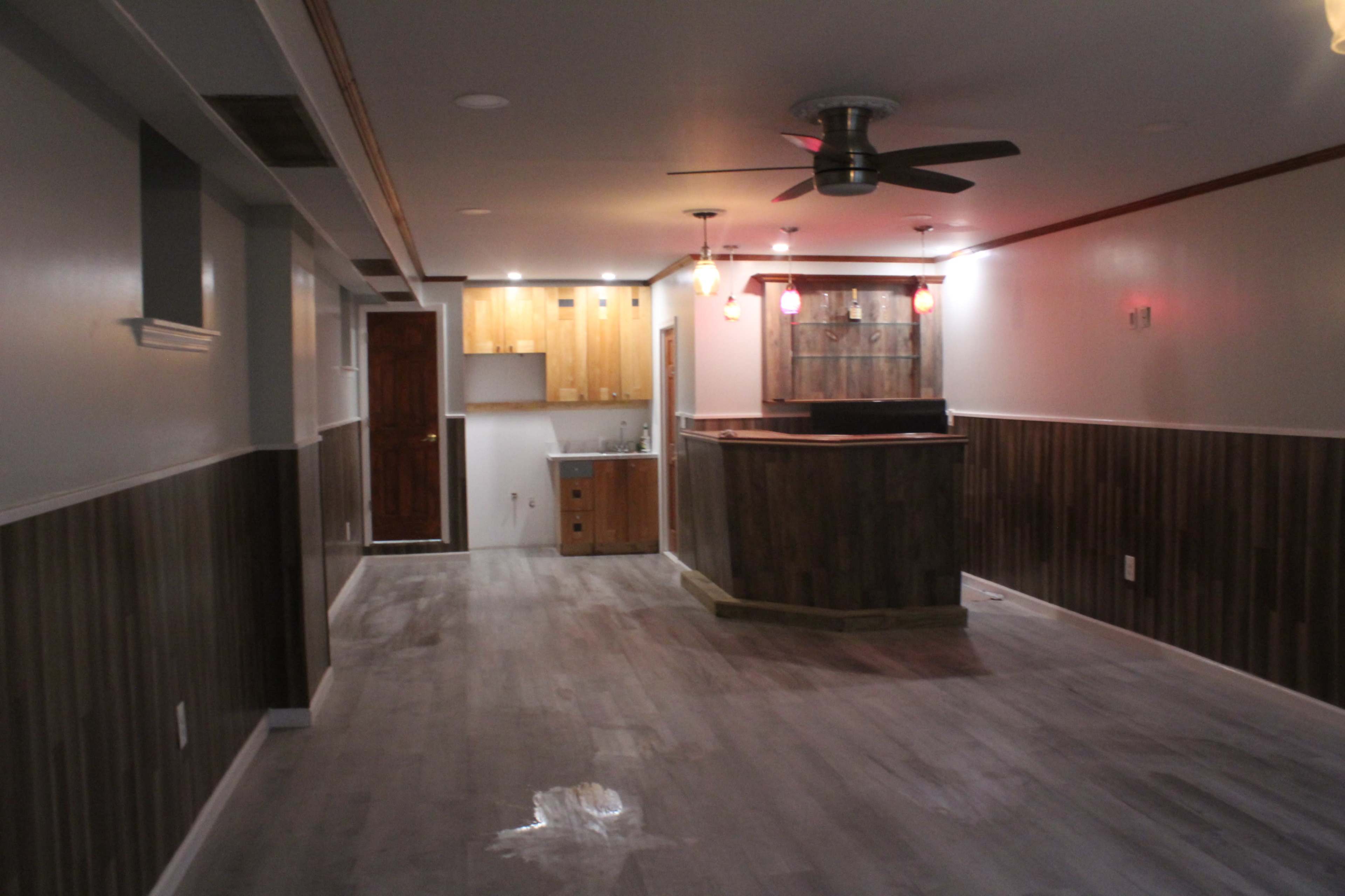 The image shows a spacious, unfinished basement area with a wooden bar, kitchenette, and a ceiling fan, featuring gray flooring and a mix of wall textures.