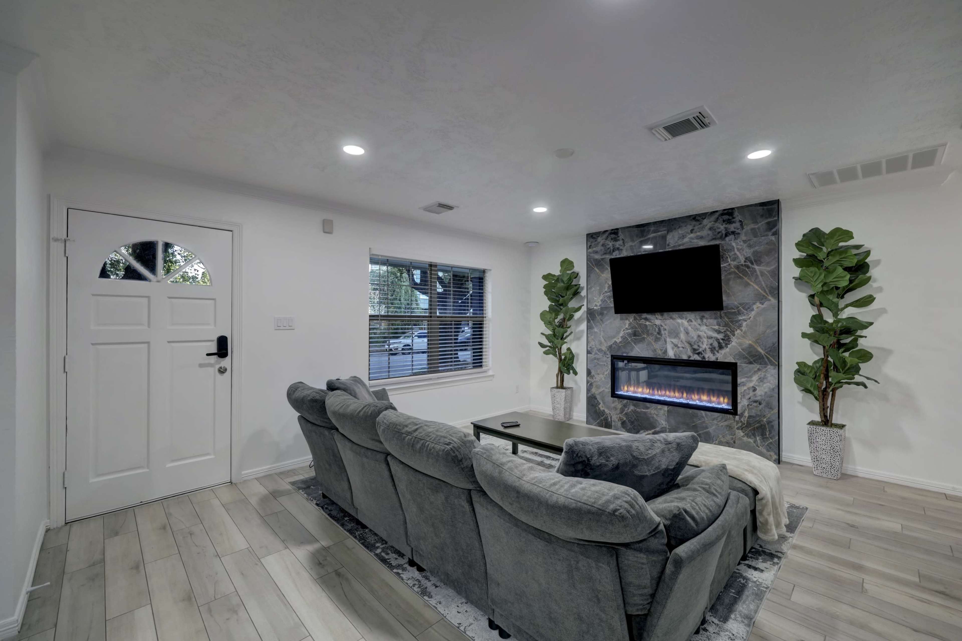 A modern living room features a gray sectional sofa, a sleek fireplace with a dark stone facade, and two tall potted plants beside a wall-mounted TV.