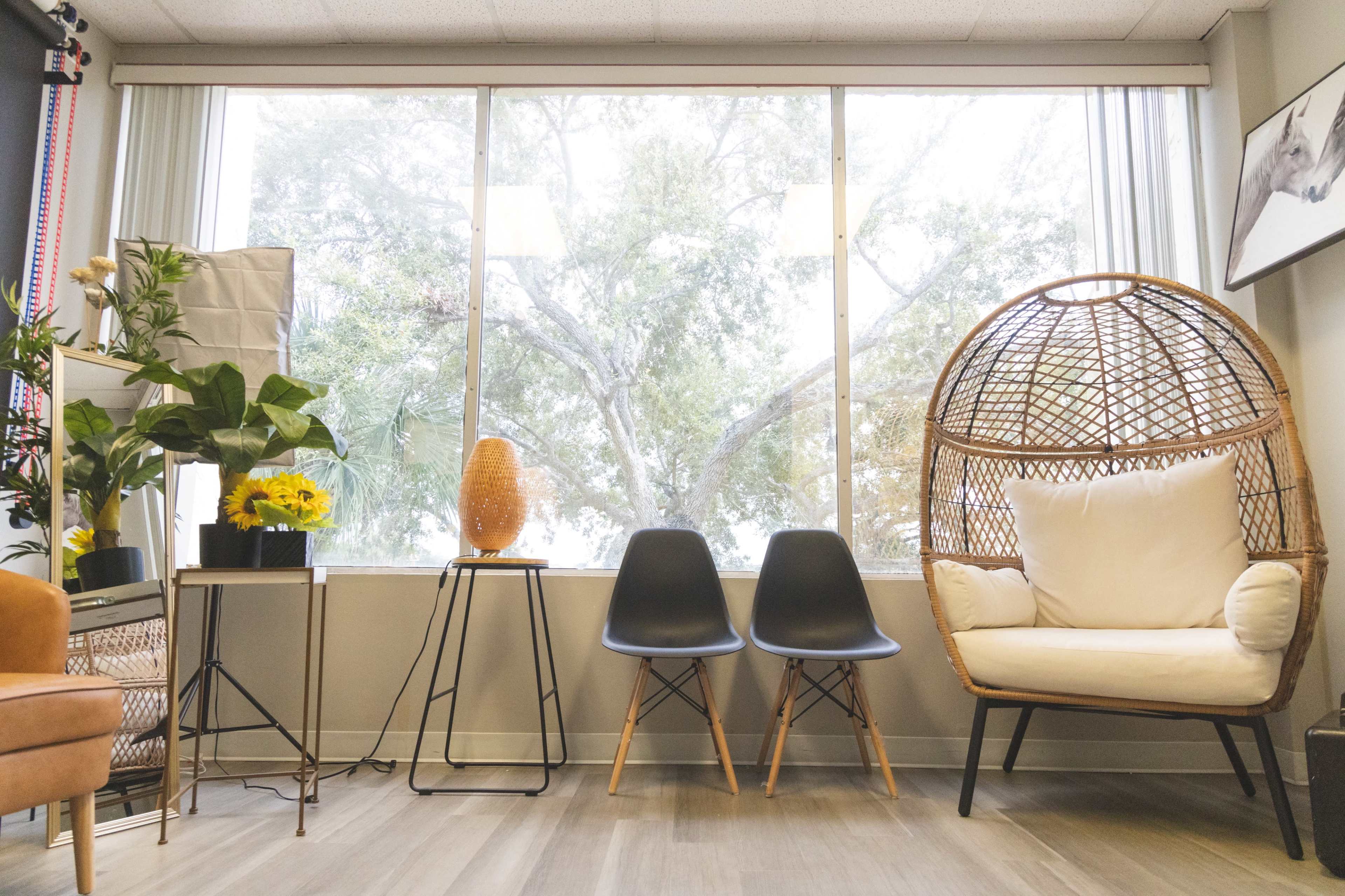 A modern living space with a large window, a rattan chair, two black chairs, a plant, and decorative lamps.