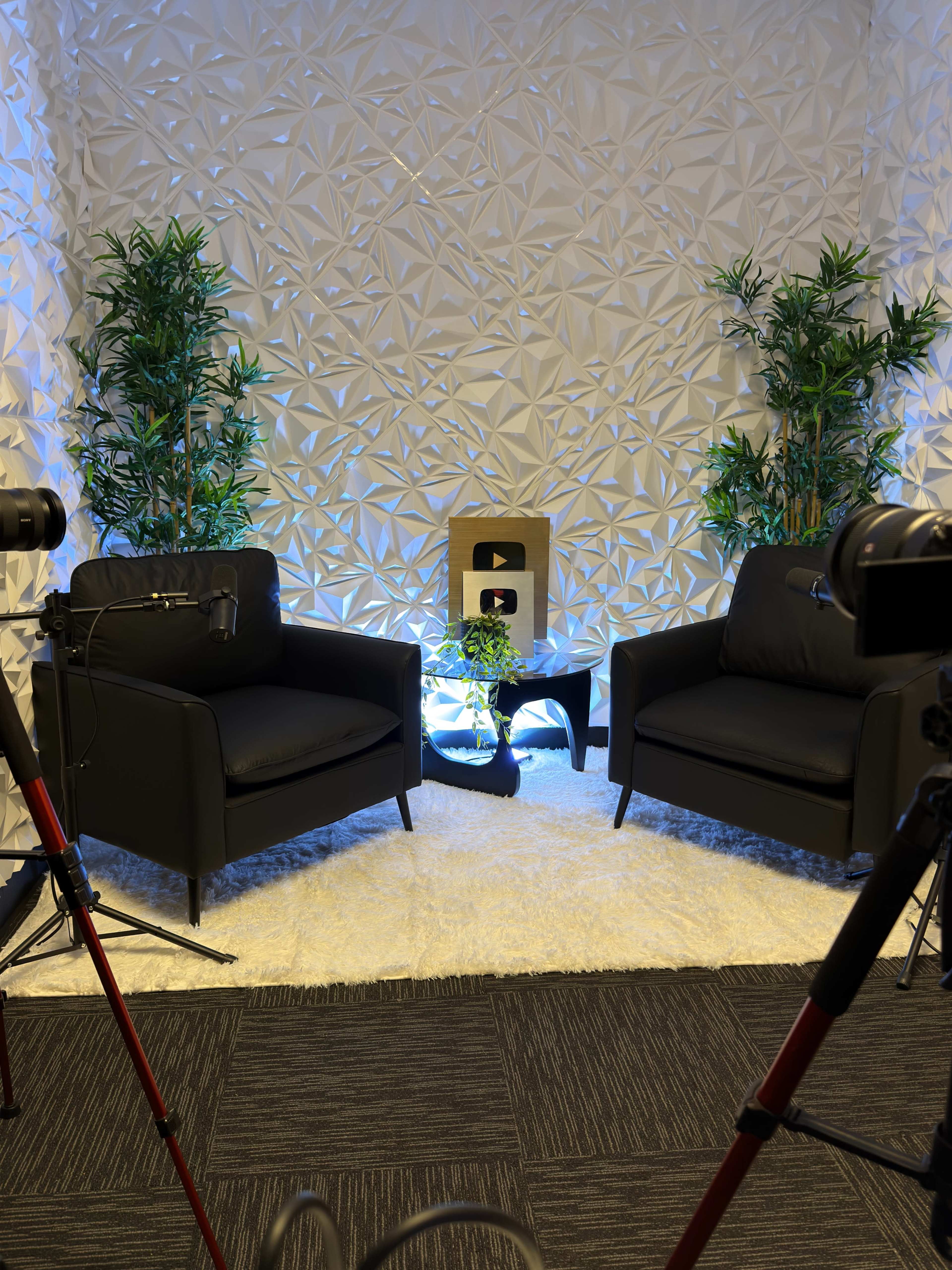 The image shows a modern interview setup with two black chairs, a decorative wall, plants, and a golden YouTube play button placed on a small table.
