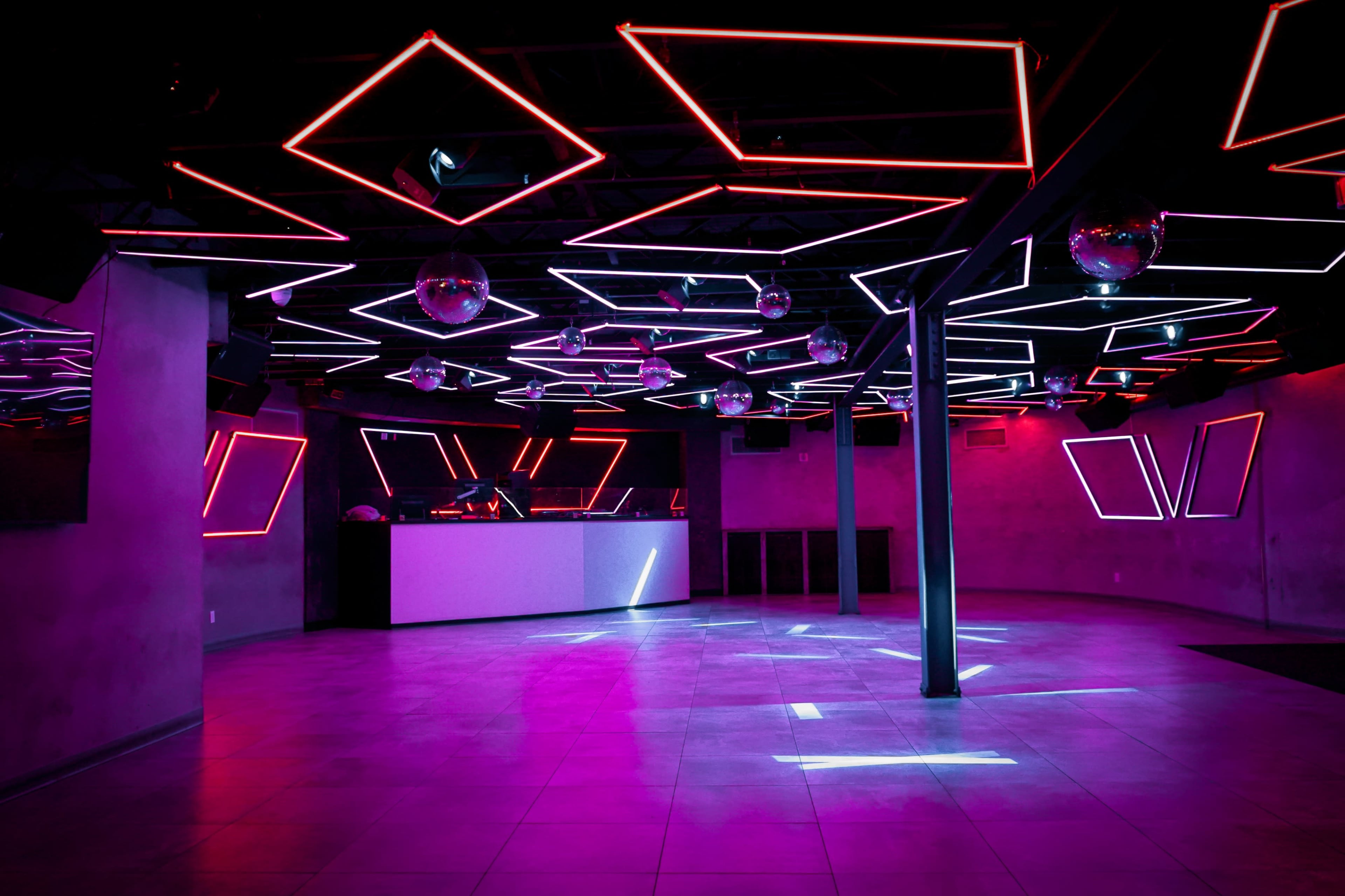 A dimly lit nightclub features a ceiling adorned with neon lights in geometric shapes and several disco balls reflecting light across the floor.