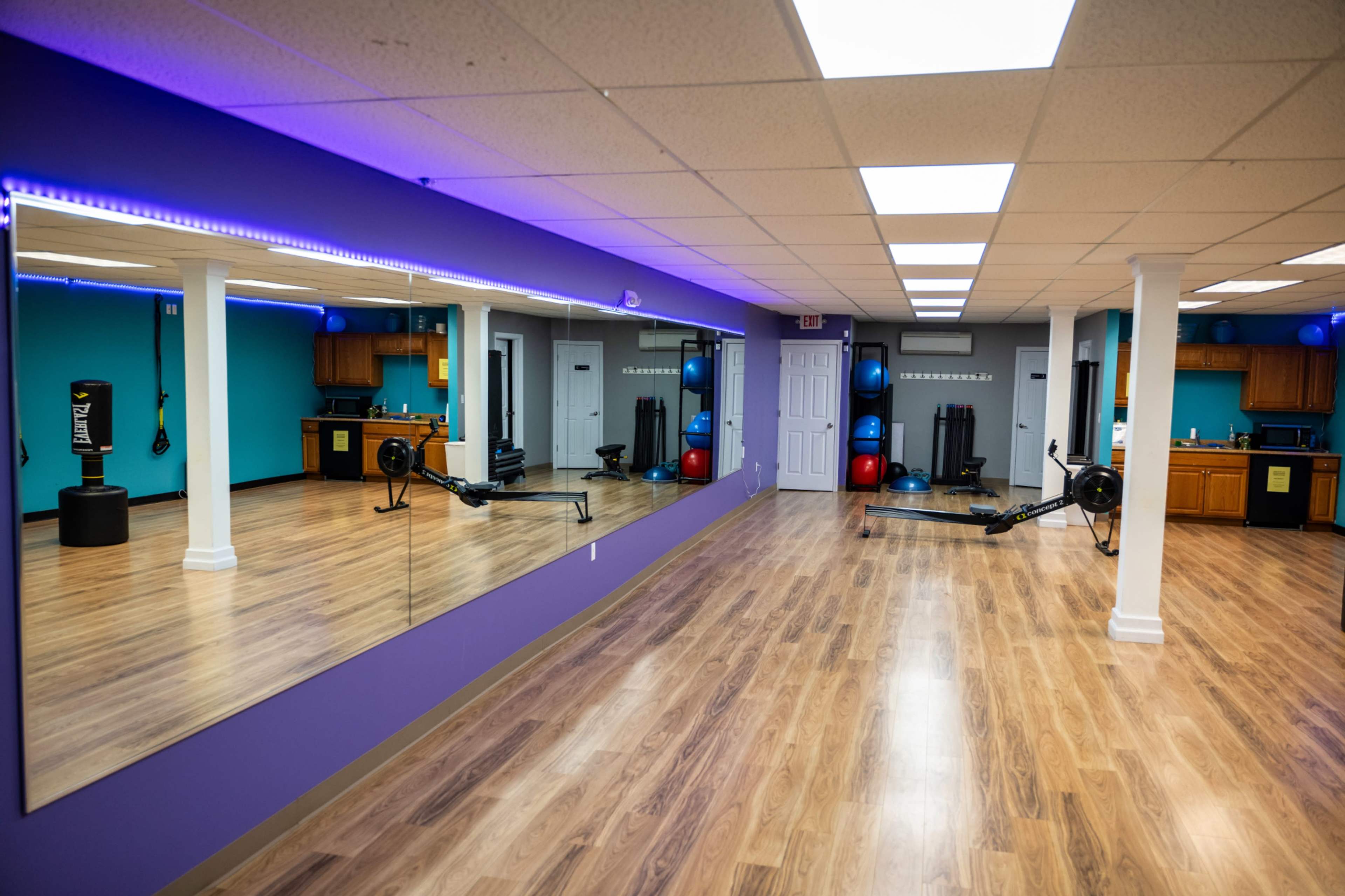 A fitness studio with wooden flooring, blue walls, mirrors, and exercise equipment including a rowing machine and a heavy bag.