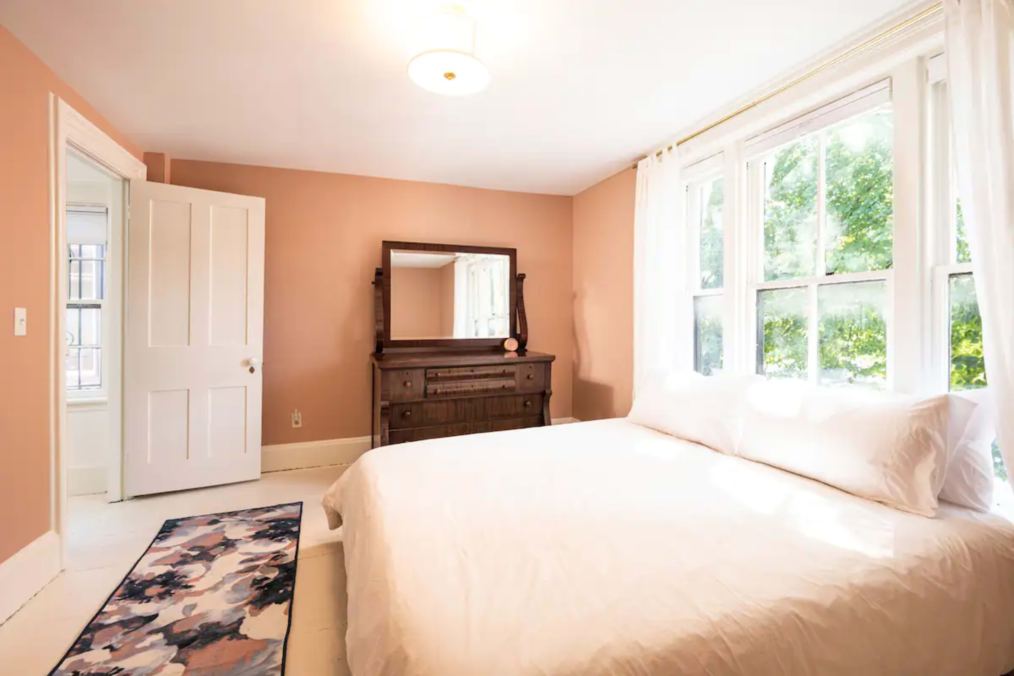 A bright bedroom featuring a white bed, a wooden dresser, and large windows allowing natural light to enter.