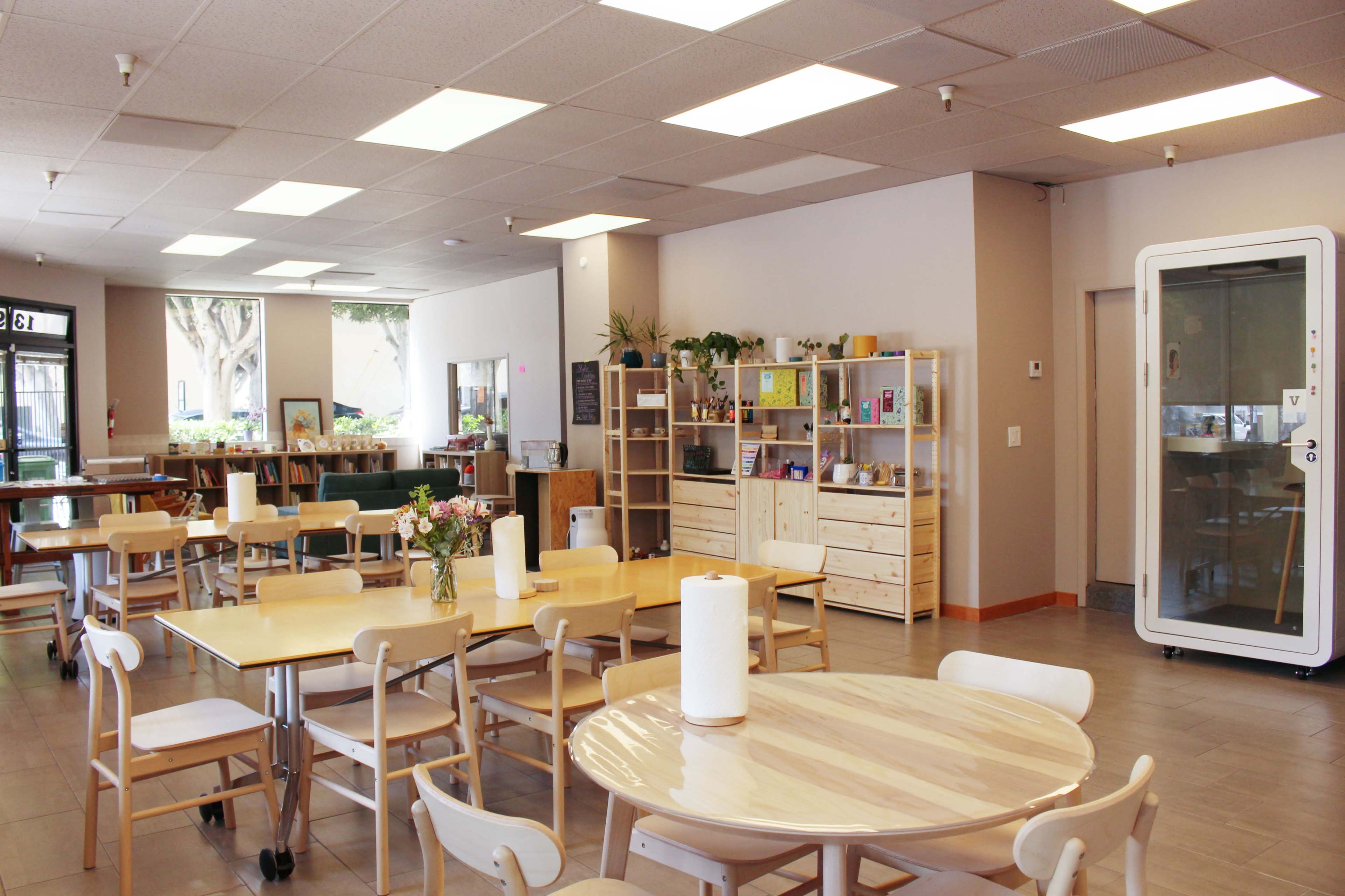 The image shows a spacious indoor area with wooden tables and chairs, shelves filled with items, and large windows letting in natural light.
