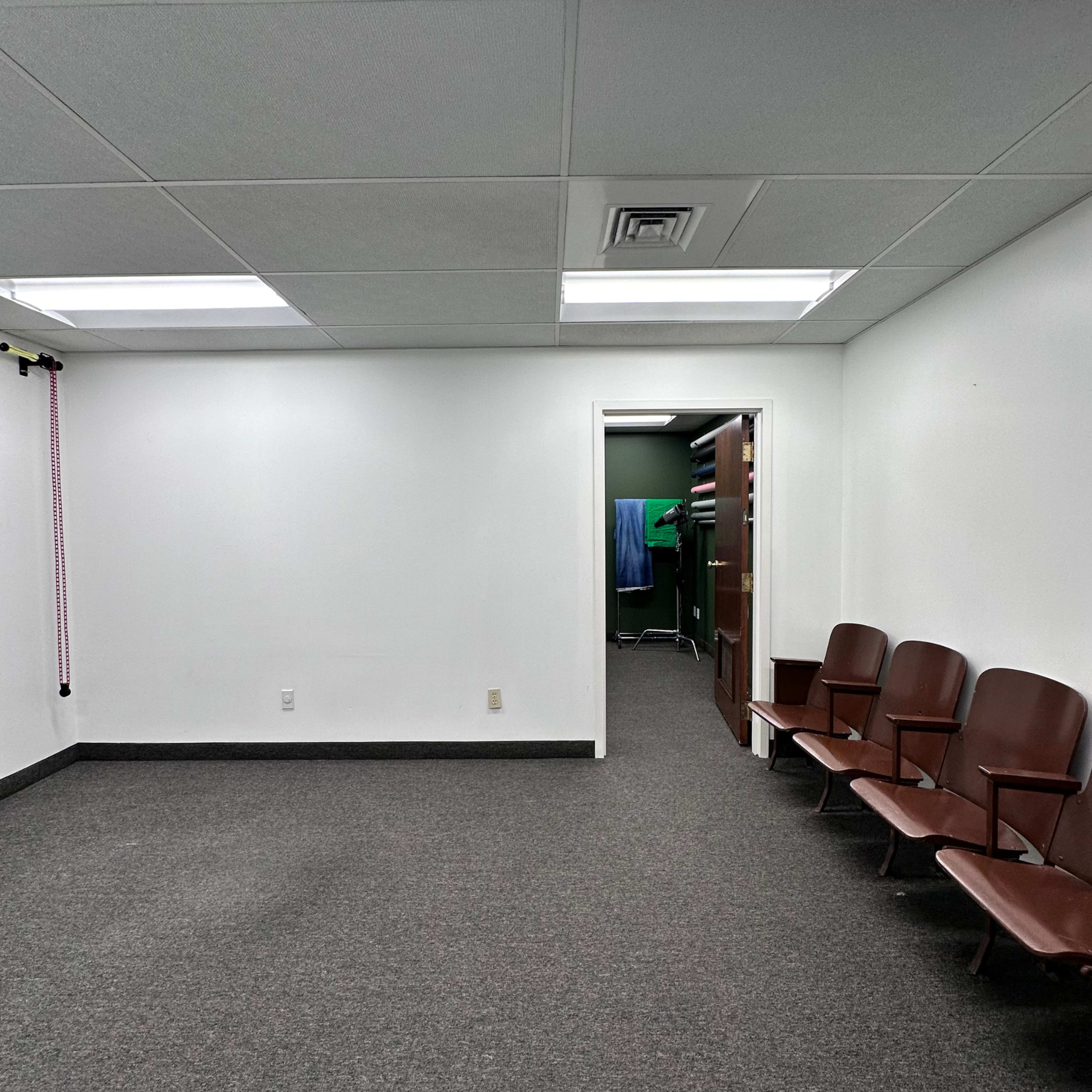 The image shows an empty room with gray carpeting, white walls, and a row of brown chairs along one side next to a door leading to a darker space.
