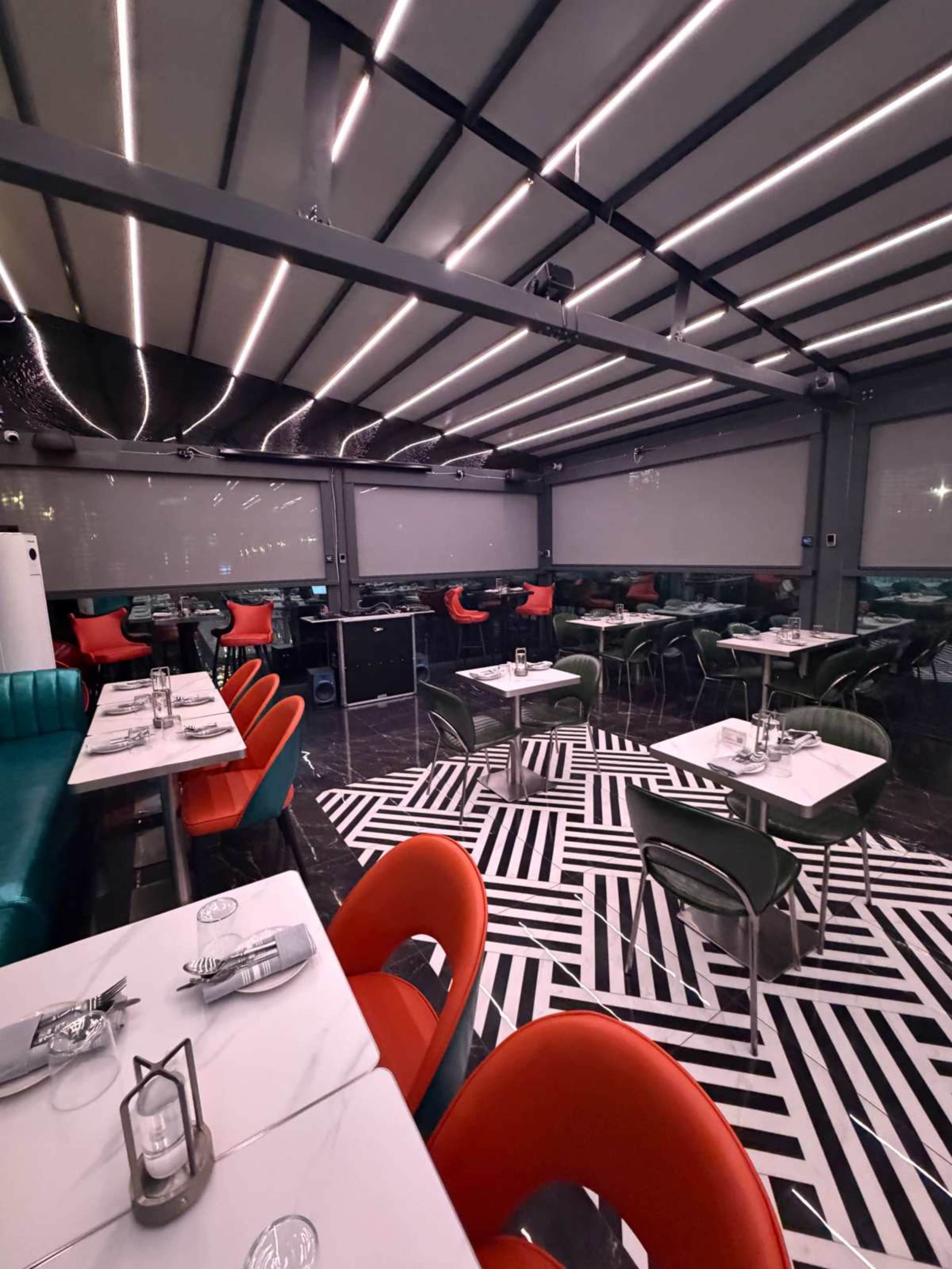 The image shows a modern restaurant interior featuring tables with white tablecloths, colorful chairs, and a striped black and white rug.