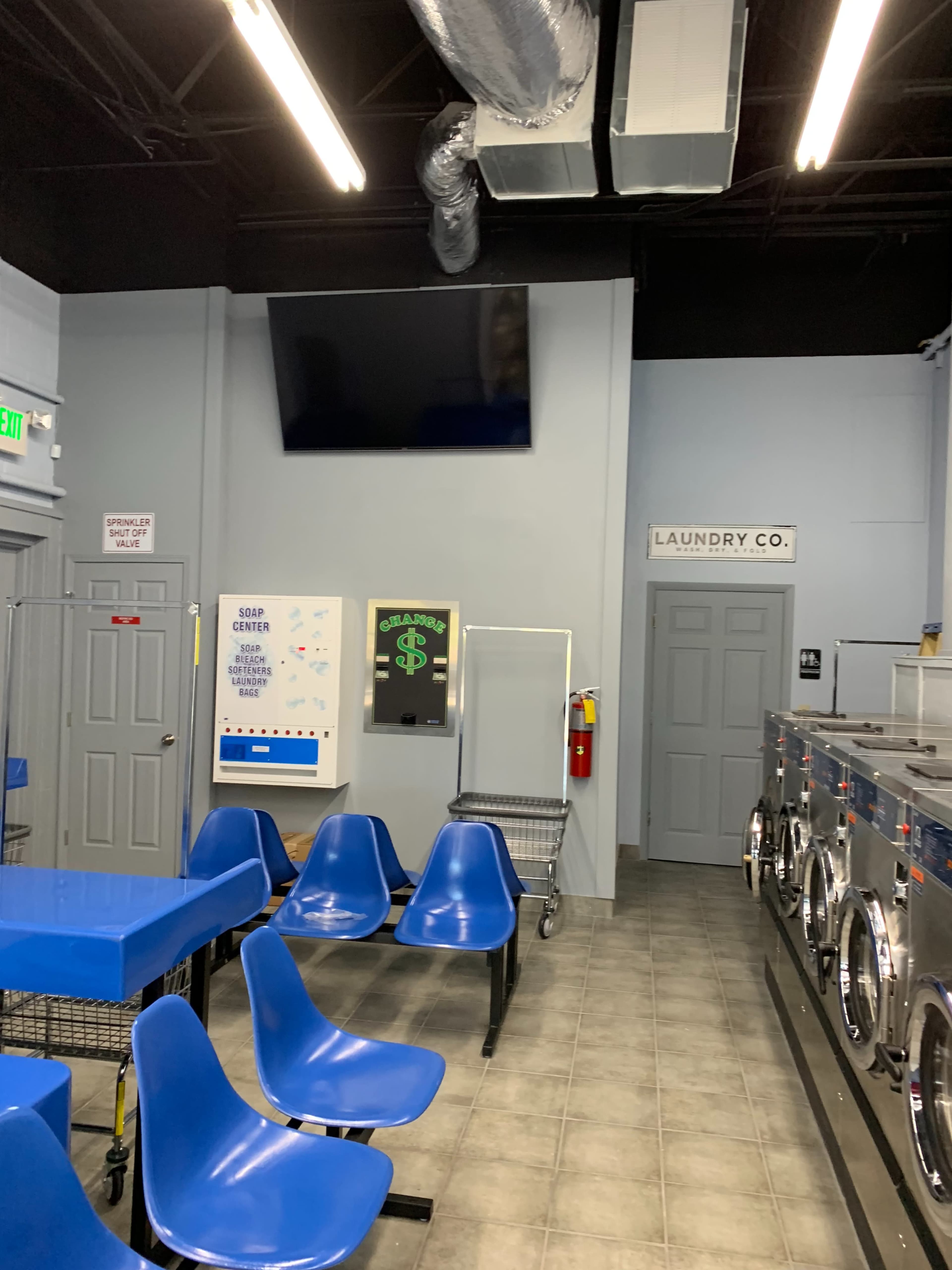 The interior of a laundry facility featuring blue plastic chairs, a wall-mounted television, and rows of washing machines.