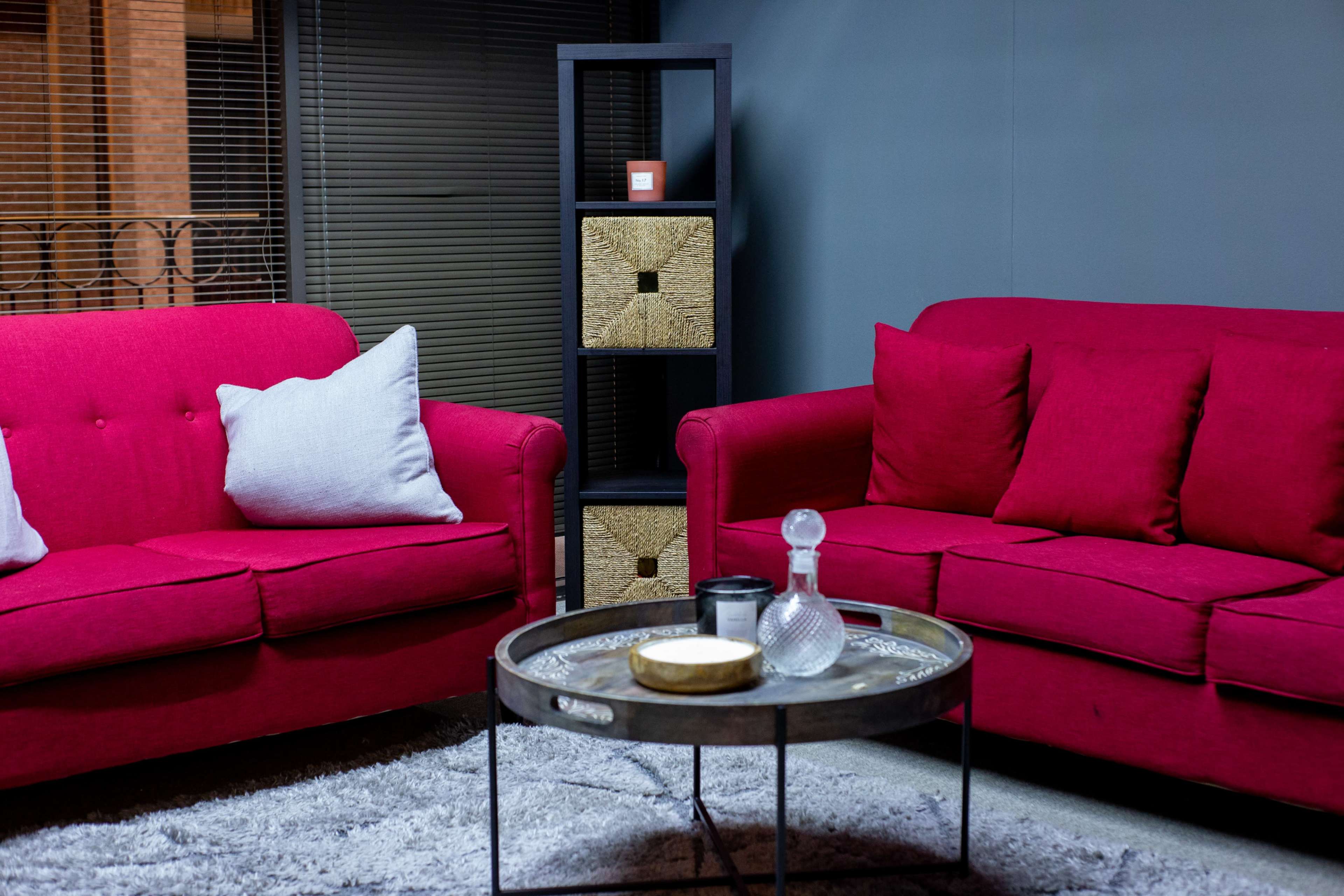 A living room features two bright pink sofas flanking a round coffee table, with a bookshelf and decorative storage boxes in the background.