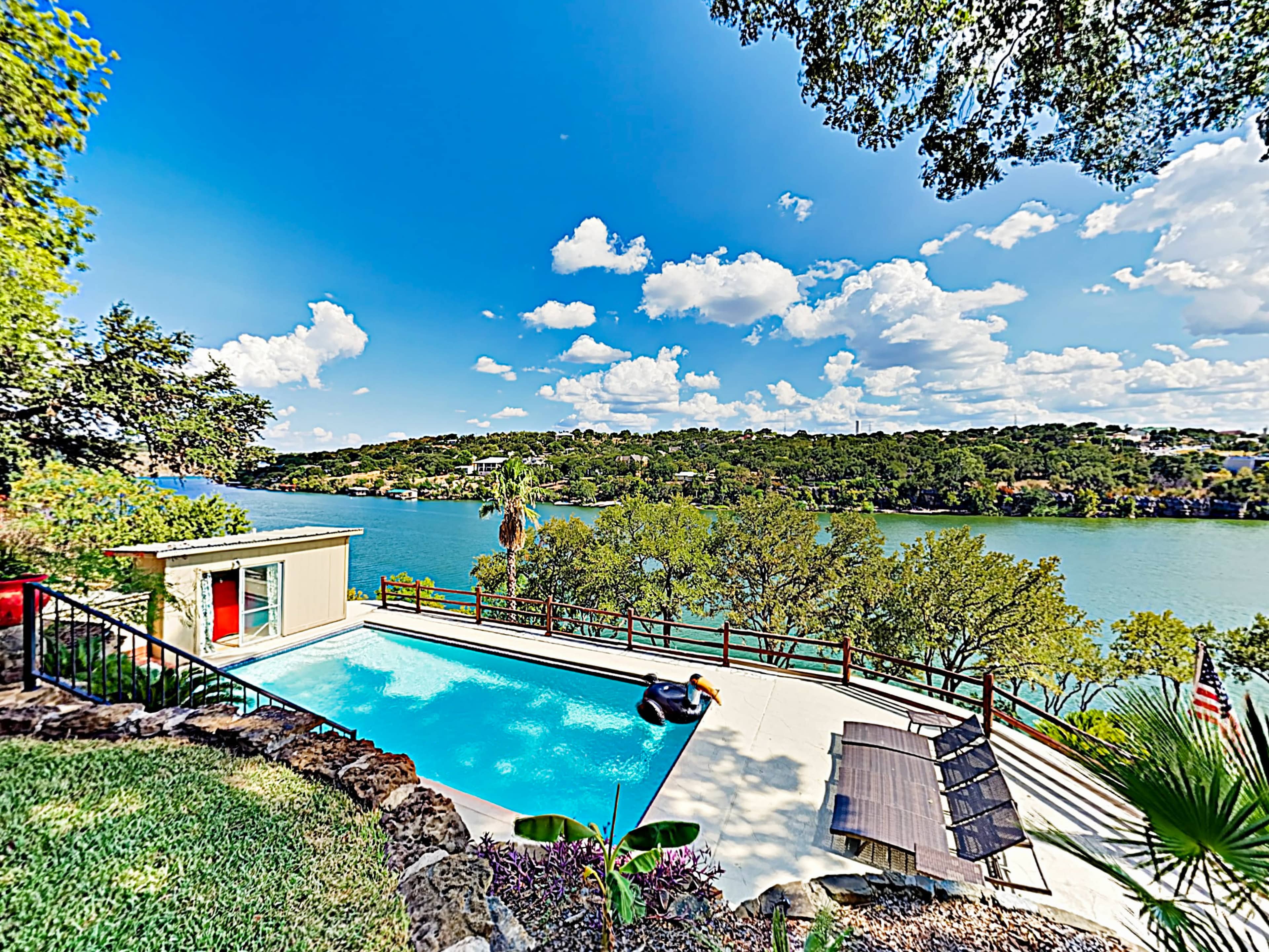 A swimming pool overlooks a river surrounded by trees and a clear blue sky filled with clouds.