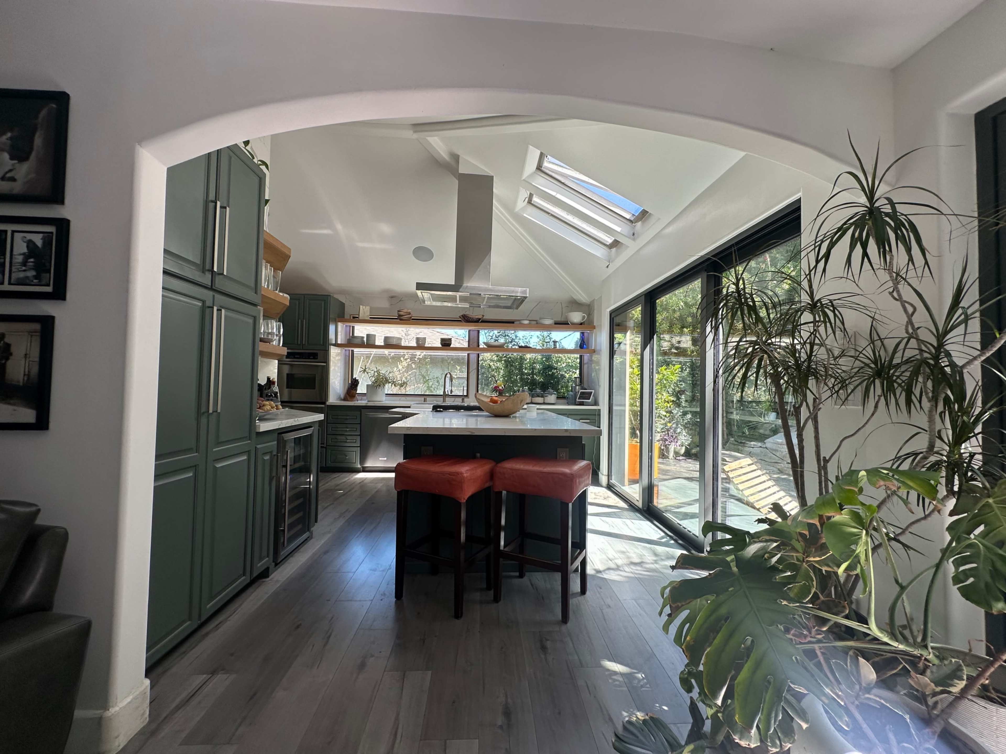 The image shows a modern kitchen featuring green cabinetry, a large island with orange bar stools, and large windows that allow natural light into the space.